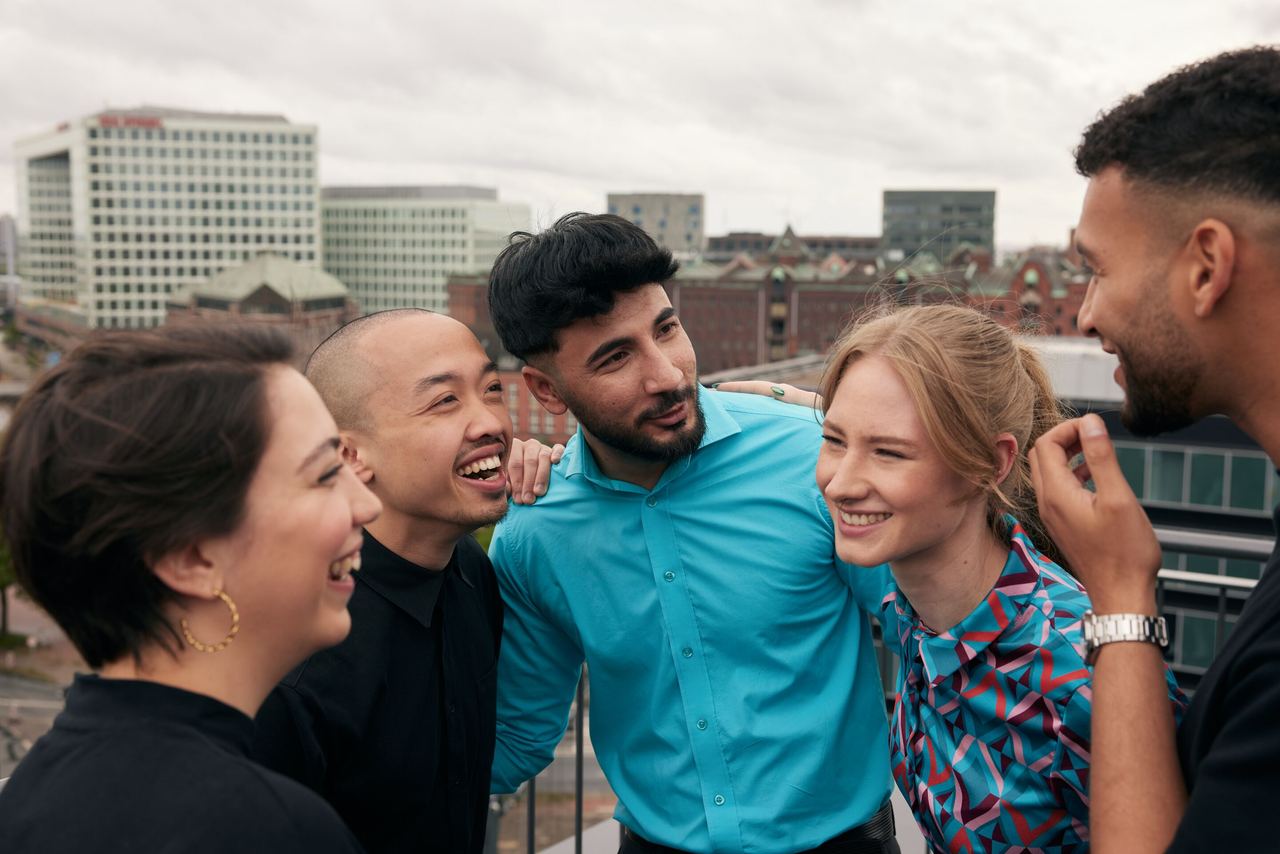 Fünf junge Menschen lachen und unterhalten sich entspannt auf einer Dachterrasse mit Blick auf die Stadt, vermittelt ein Gefühl von Gemeinschaft und guter Stimmung.