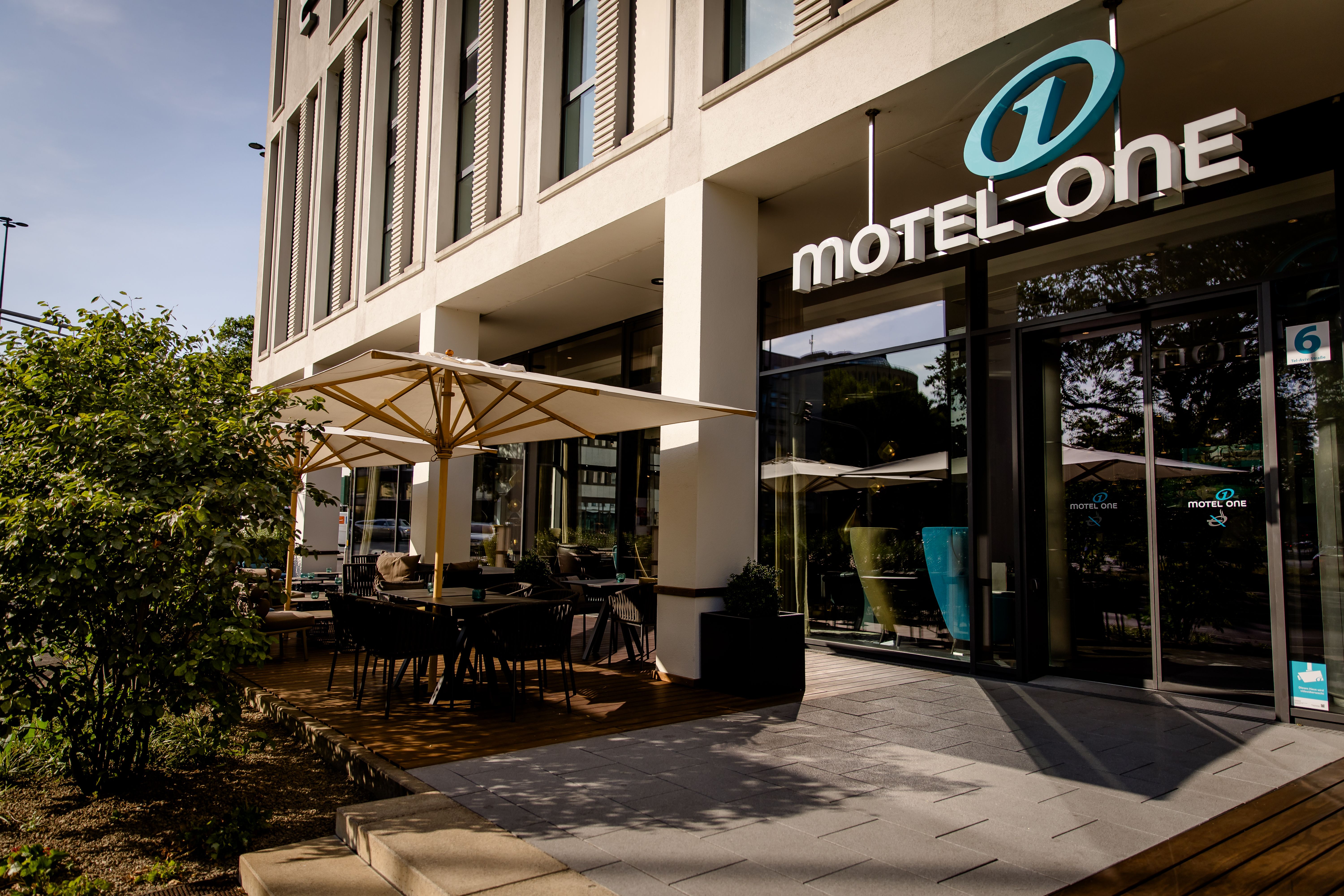 Eingangsbereich des Motel One mit überdachter Außenterrasse, Stühlen und Tischen, moderner Fassade und großem Logo, ideal für Gäste, die Komfort und stilvolles Ambiente suchen.