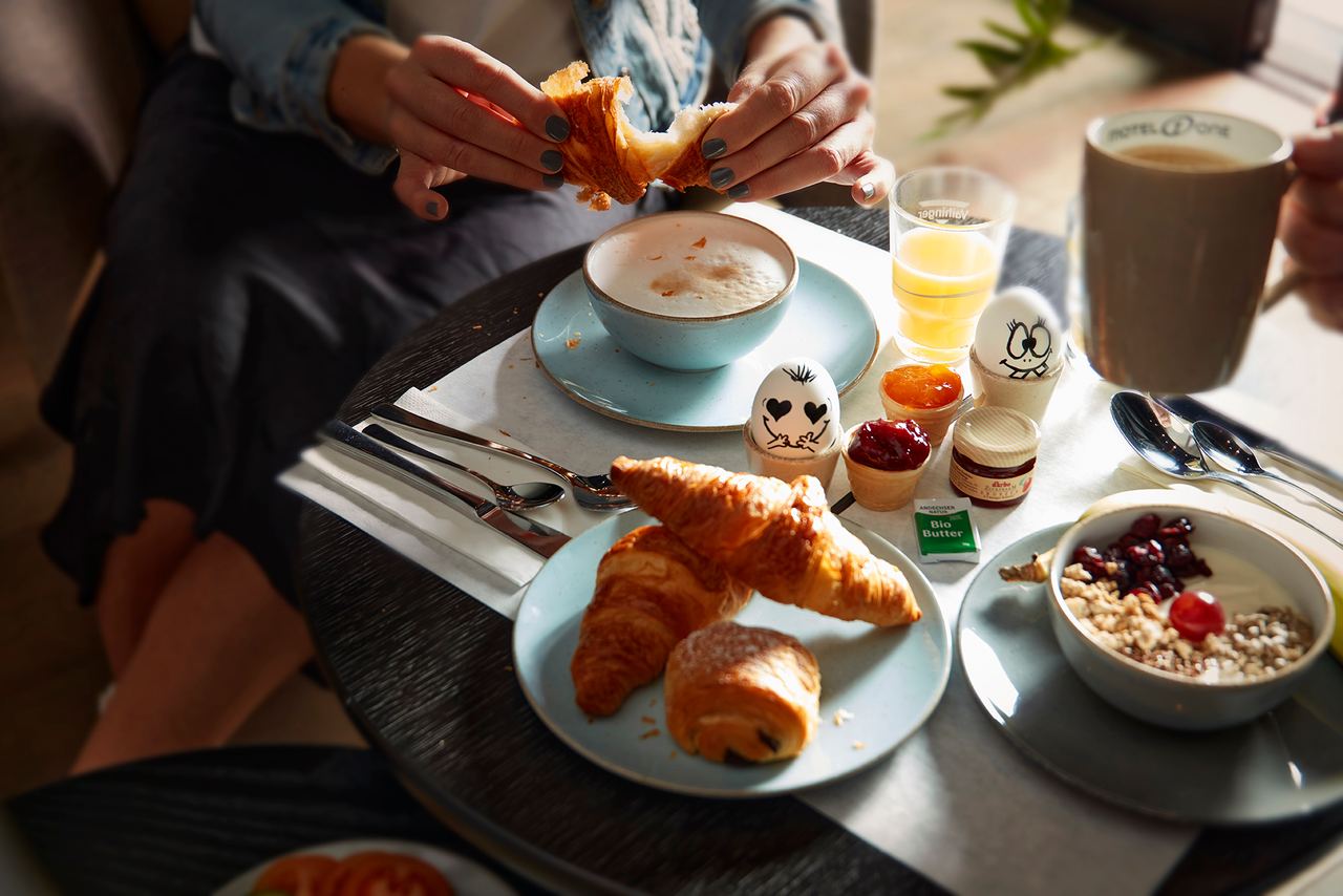 Person genießt entspannt Frühstück mit Croissants, Cappuccino, Müsli, frisch gepresstem Saft und fruchtigen Aufstrichen in gemütlicher Atmosphäre.