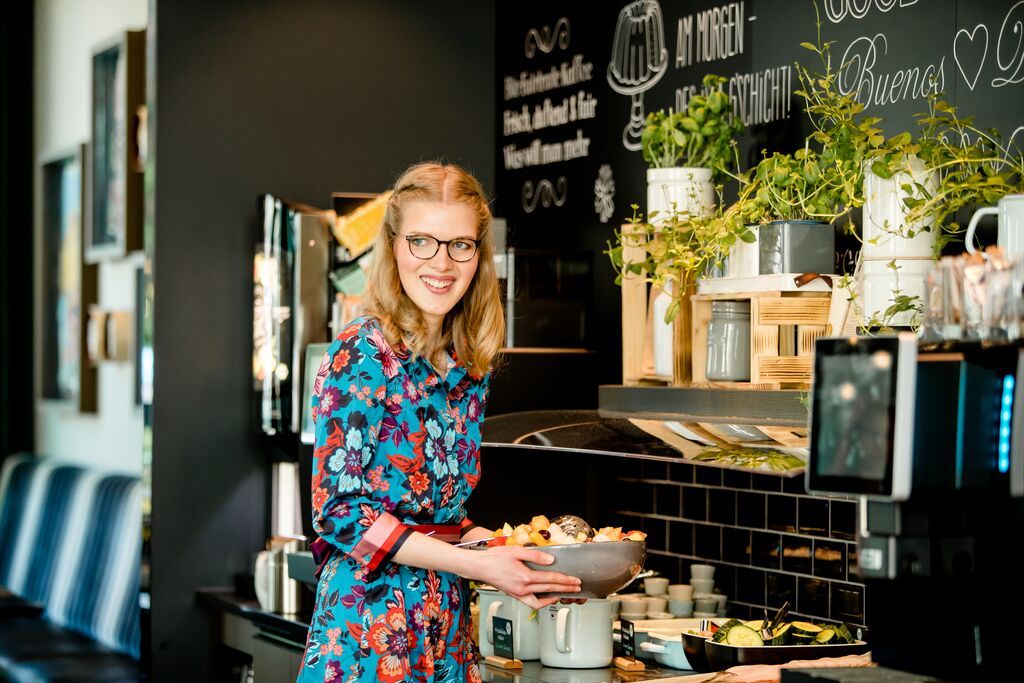 Frau mit Brille und buntem Kleid steht lächelnd an einem Buffet mit frischem Obst und Pflanzen, die Atmosphäre wirkt einladend und gemütlich.