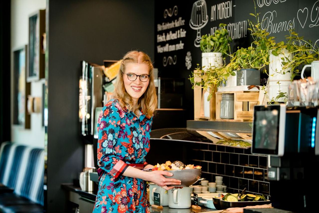 Frau mit Brille und buntem Kleid steht lächelnd an einem Buffet mit frischem Obst und Pflanzen, die Atmosphäre wirkt einladend und gemütlich.