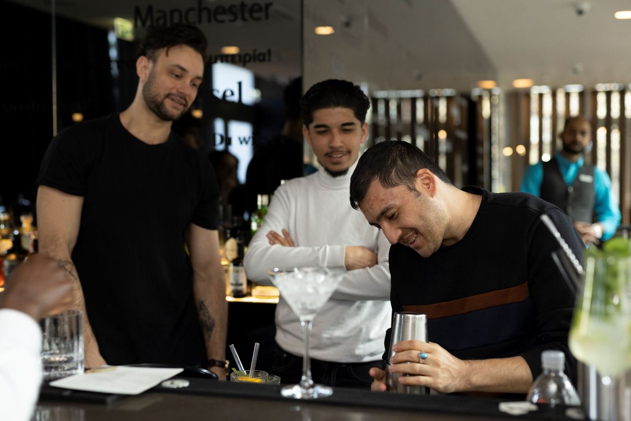 Drei Männer beobachten interessiert einen Barkeeper, der einen Cocktail an der Hotelbar zubereitet. Die Atmosphäre wirkt entspannt und gesellig.