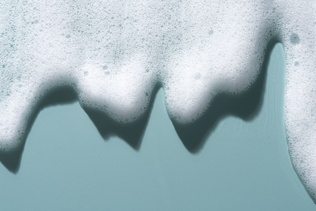 Schaum von Seifenblasen auf einer glatten, hellblauen Oberfläche, der sanft in unregelmäßigen Formen verläuft und eine ruhige, entspannende Atmosphäre erzeugt.