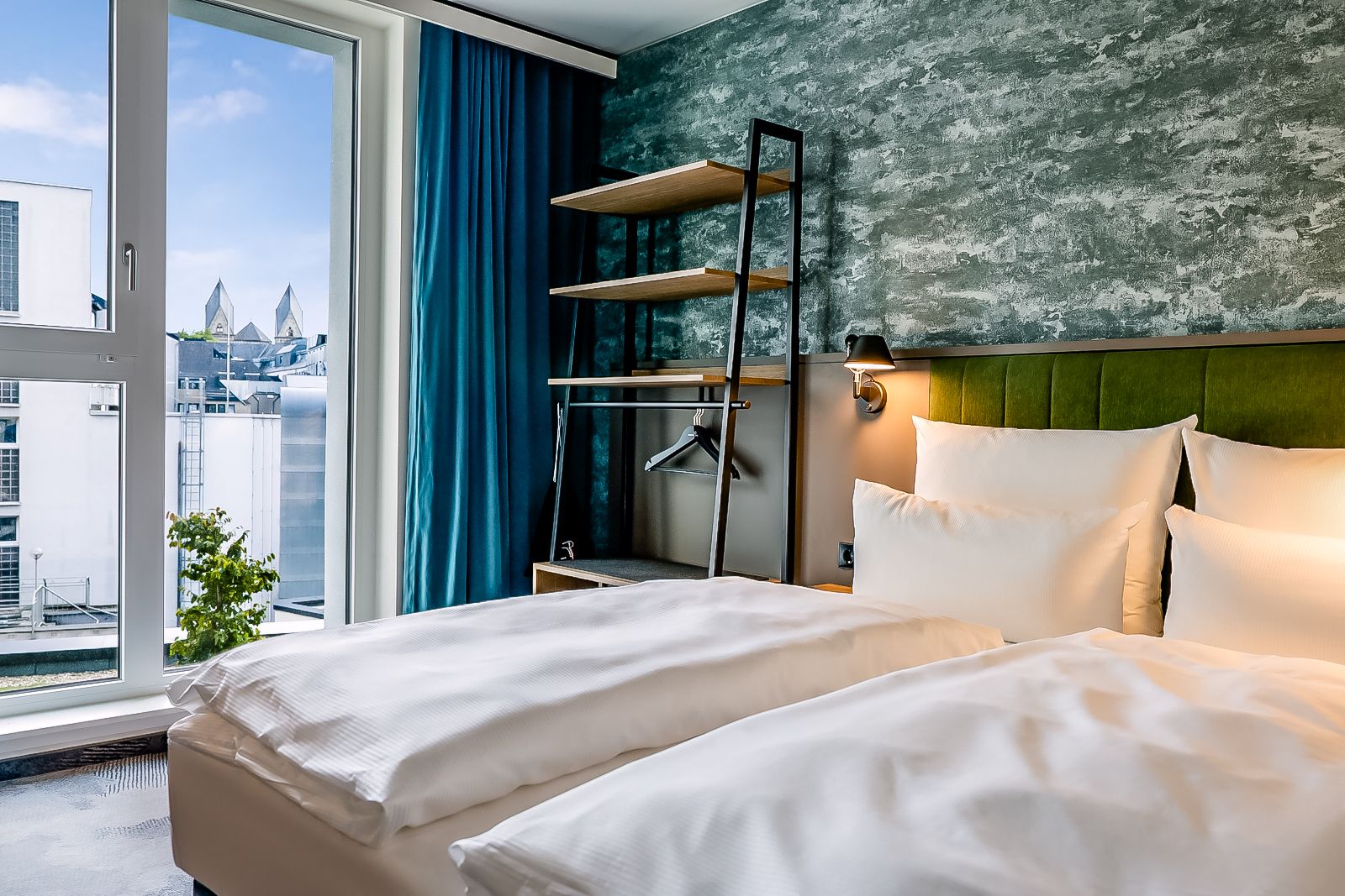 Gemütliches Hotelzimmer mit zwei Einzelbetten, weißen Bettwäsche, einer grünen gepolsterten Kopfteilwand, offener Garderobe und großem Fenster mit Blick auf Stadtgebäude und blauen Himmel. Moderne, elegante Atmosphäre, ideal für erholsamen Aufenthalt.
