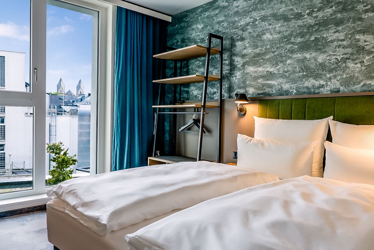 Gemütliches Hotelzimmer mit zwei Einzelbetten, weißen Bettwäsche, einer grünen gepolsterten Kopfteilwand, offener Garderobe und großem Fenster mit Blick auf Stadtgebäude und blauen Himmel. Moderne, elegante Atmosphäre, ideal für erholsamen Aufenthalt.