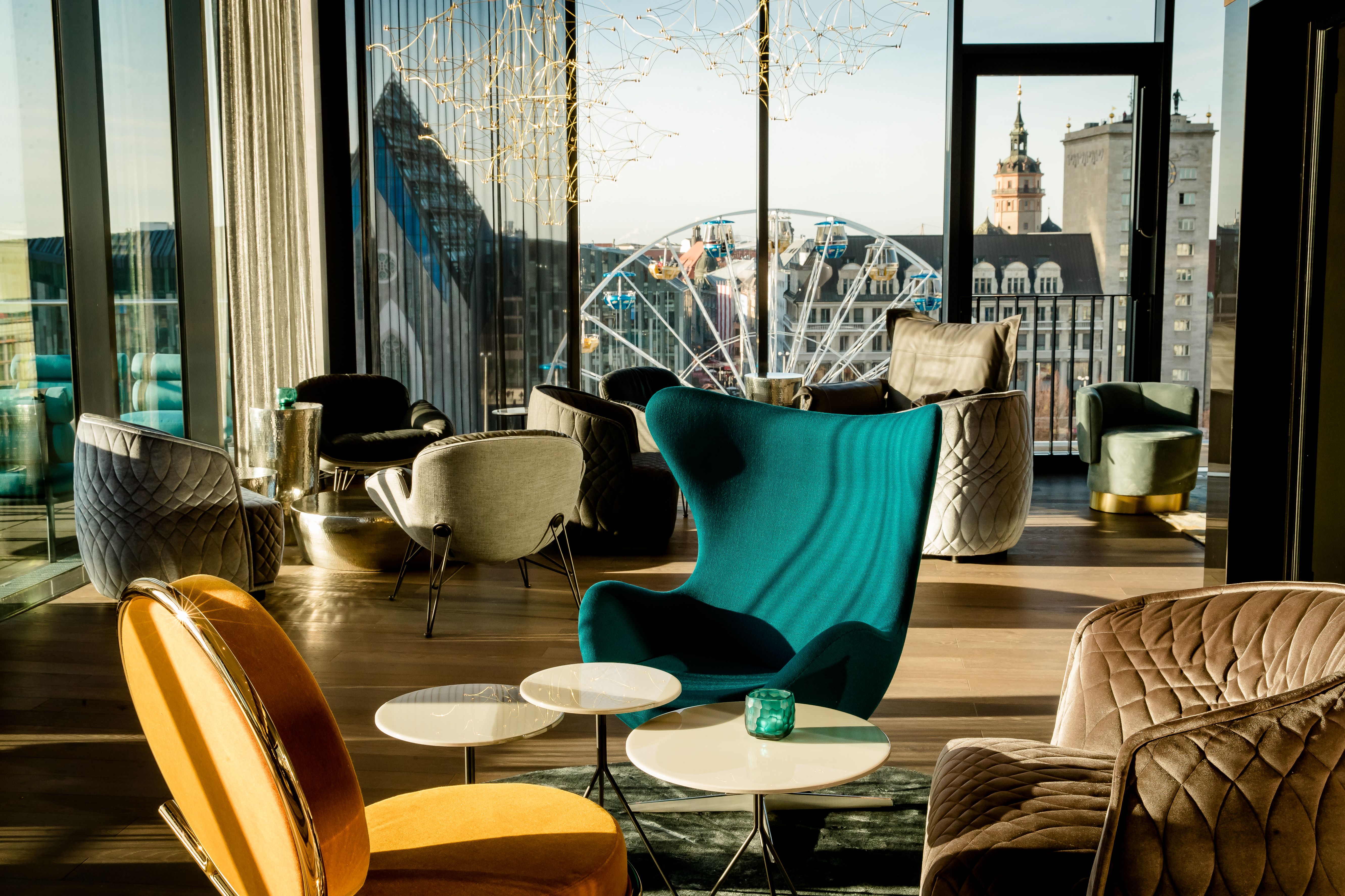 Moderne Lounge mit gemütlichen, farbenfrohen Sesseln vor großen Fenstern, die eine Stadtansicht mit einem Riesenrad und historischen Gebäuden zeigen. Helle Atmosphäre durch Tageslicht, stilvolle Einrichtung zum Entspannen und Genießen.