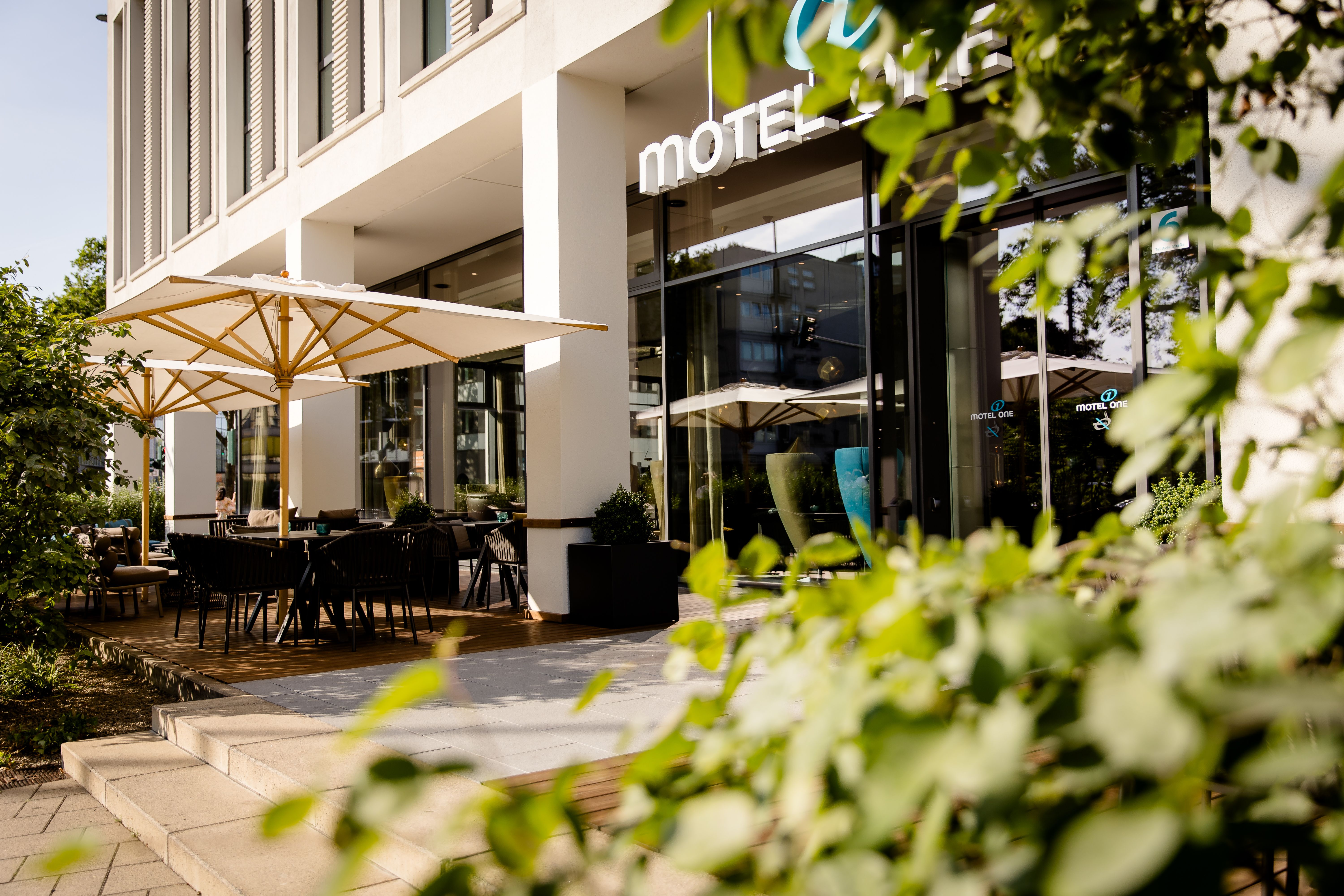 Moderner Hotel-Eingangsbereich mit Terrasse, Tischen und Sonnenschirmen, umgeben von grünen Pflanzen, einladend für Gäste zum Entspannen und Verweilen im Freien.
