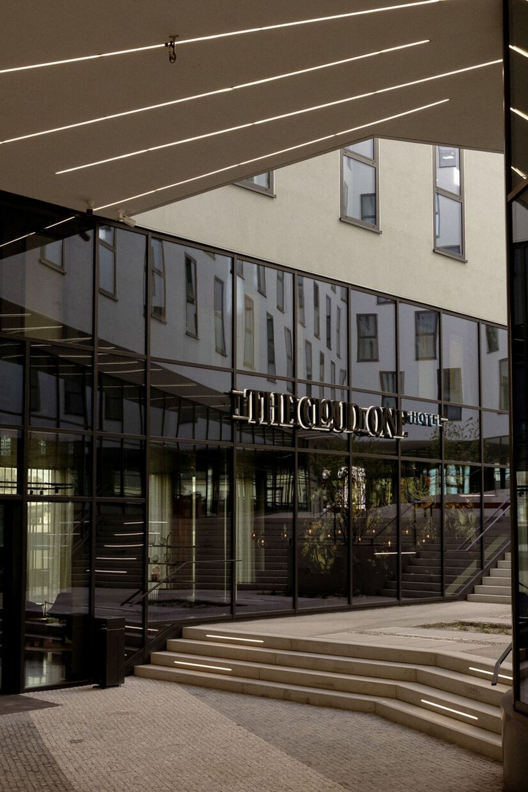 Hoteleingang mit Glasfassade und großzügiger, heller Lobby, stufenförmig angelegtem Zugang und reflektierenden Oberflächen