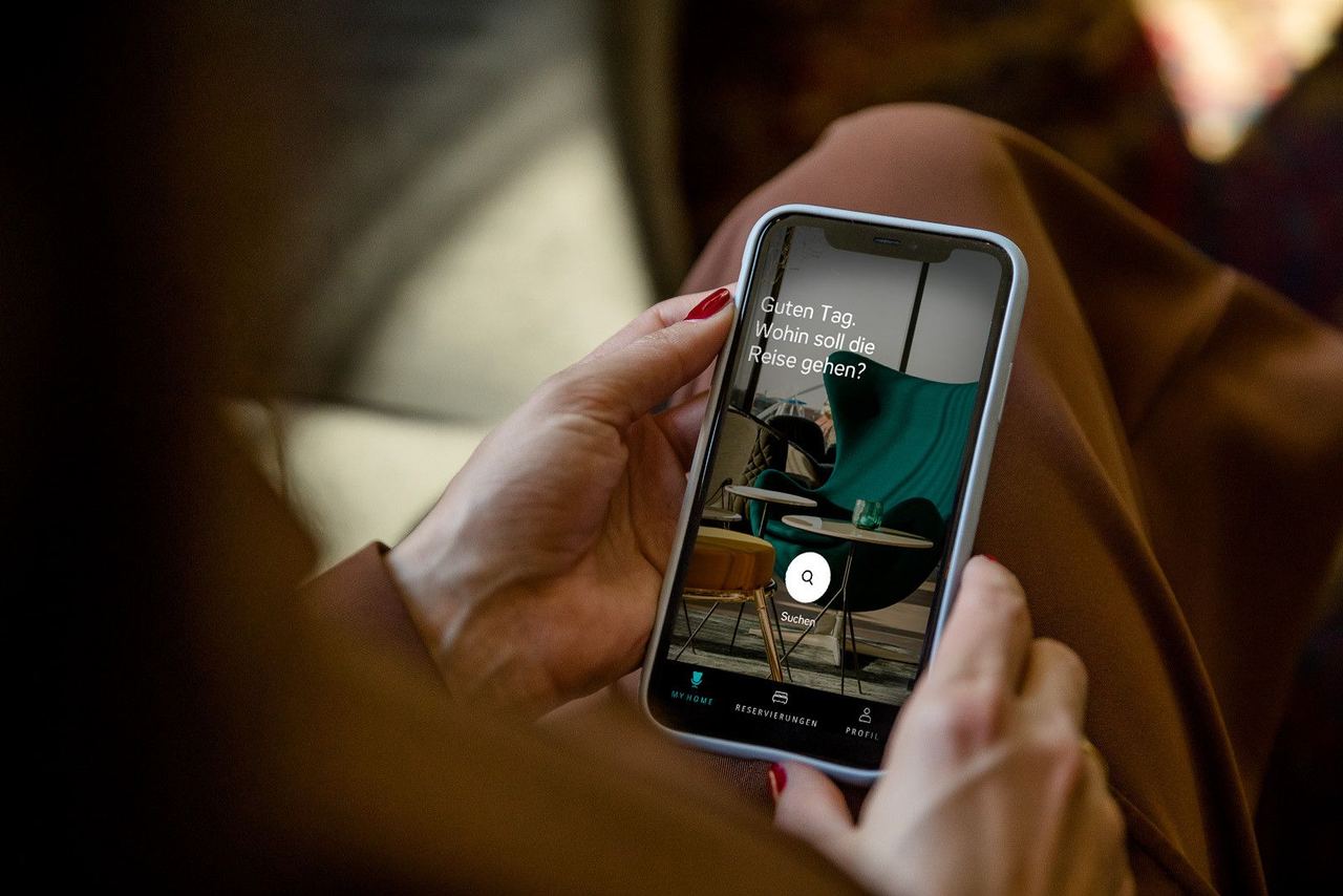 Person hält ein Smartphone, auf dem eine Hotel- oder Reise-App mit dem Text „Guten Tag, wohin soll die Reise gehen?“ angezeigt wird. Die App zeigt ein stilvolles Café-Ambiente im Hintergrund.