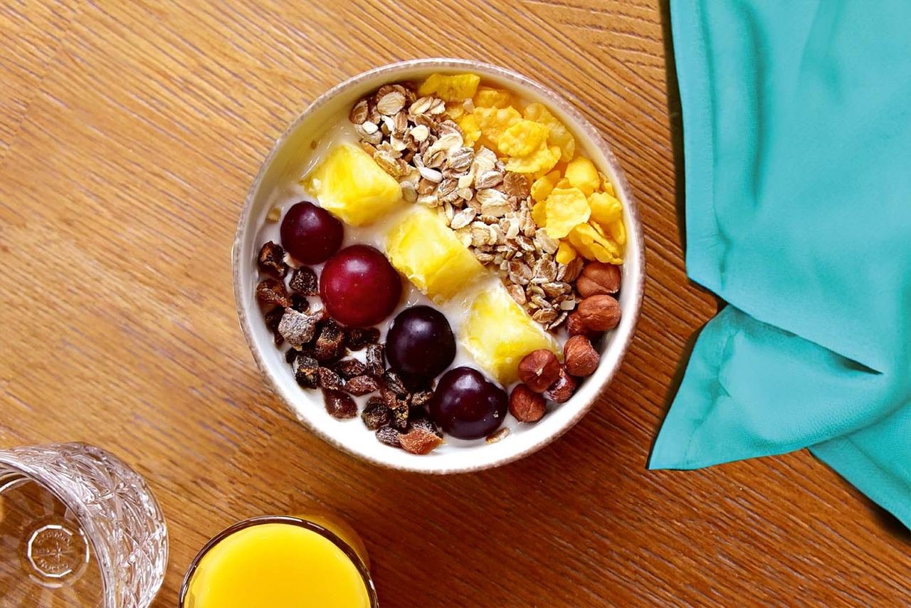 Schüssel mit Joghurt, garniert mit Trauben, Ananasstücken, Haferflocken, Cornflakes und Haselnüssen, auf einem Holztisch, neben Glas Orangensaft und Wasser.