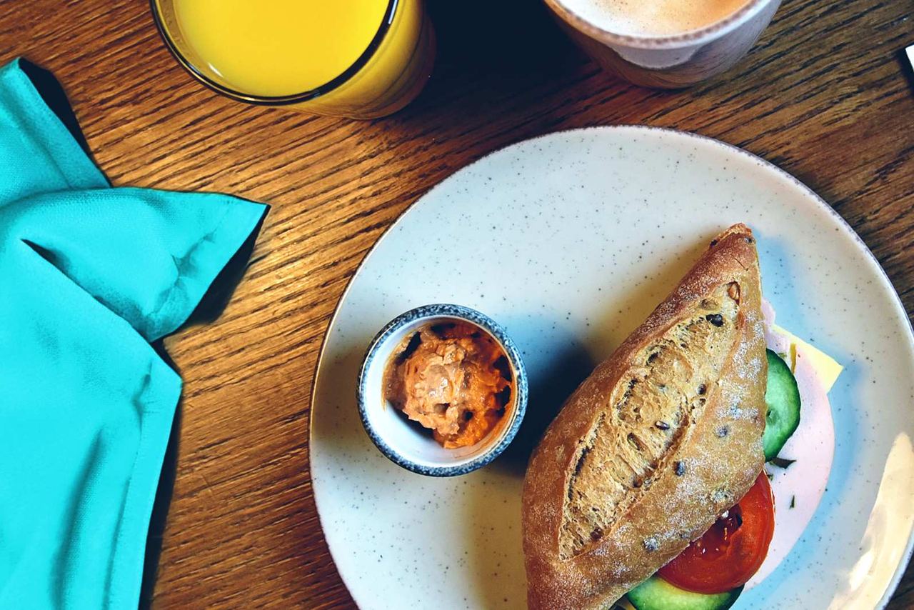 Frühstücksteller mit frischem Vollkornbrötchen, Tomaten- und Gurkenscheiben, einem kleinen Becher mit Aufstrich, dazu ein Glas Orangensaft und eine Tasse Kaffee auf Holztisch.