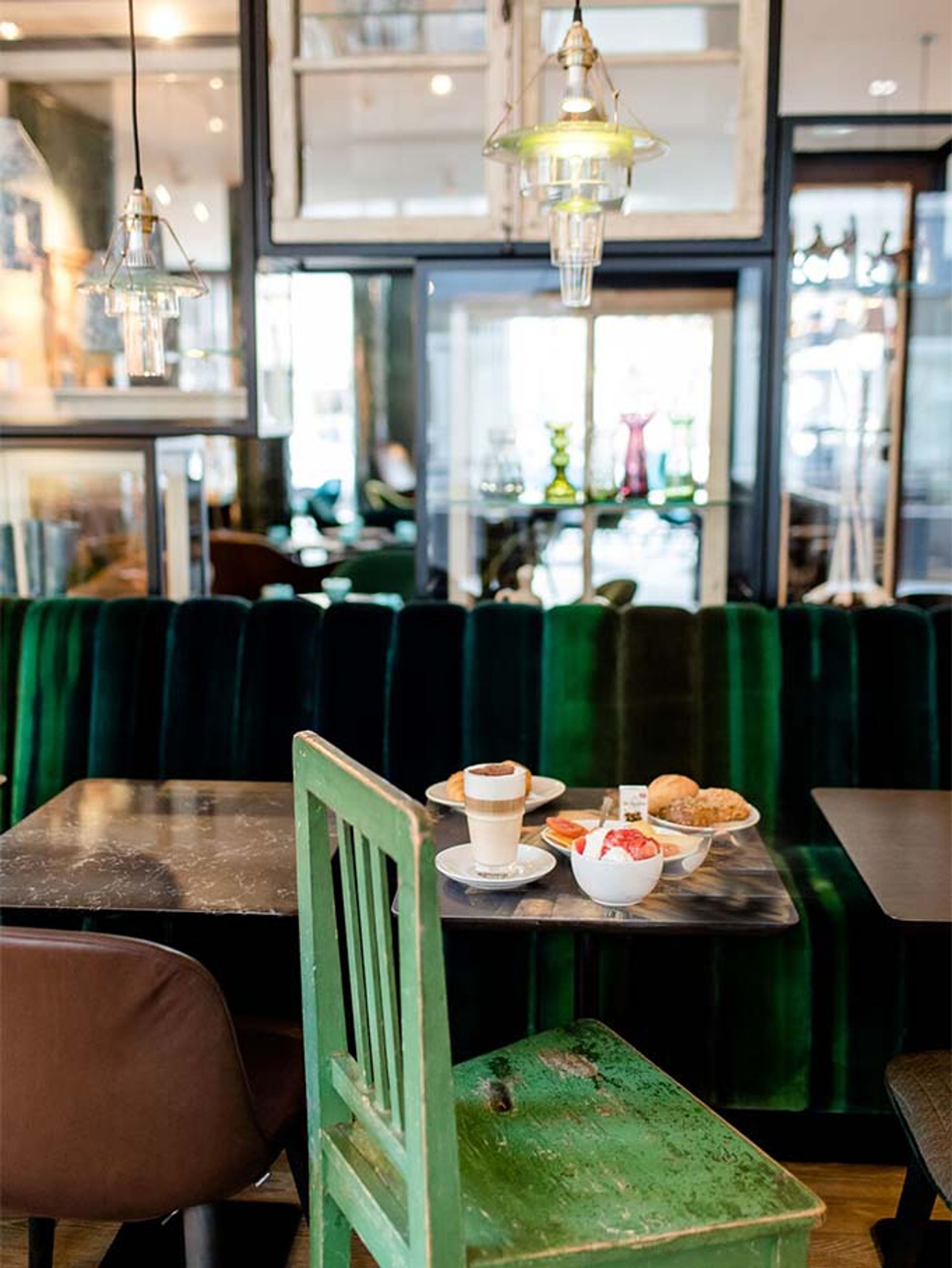 Frühstückstisch in gemütlichem Café mit Latte Macchiato, Joghurt mit Früchten und Gebäck, umgeben von grünen Polstersesseln und warmem Licht.
