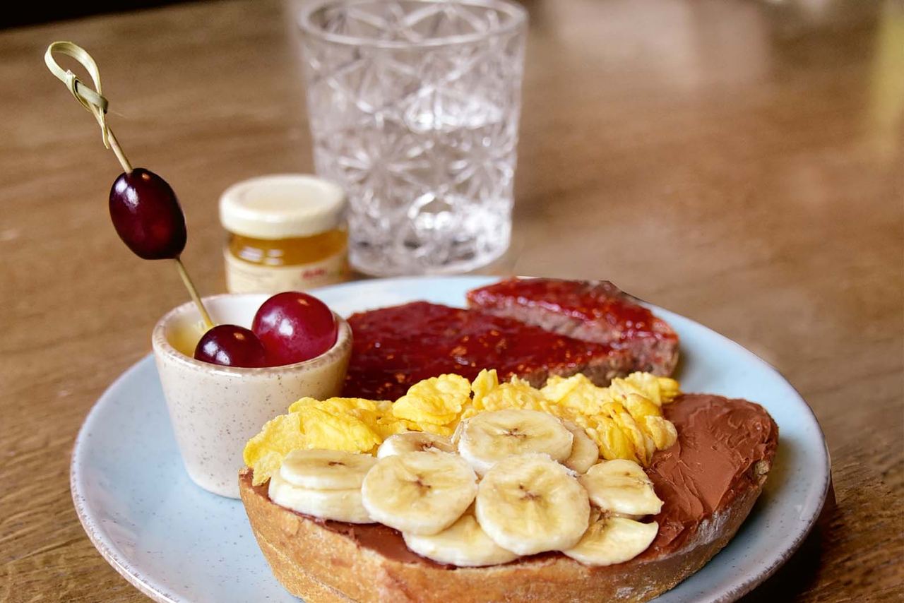 Frühstücksteller mit einem Toast, bestrichen mit Schokolade, belegt mit Bananenscheiben, dazu Rührei, eine Scheibe Wurst und ein kleiner Becher mit Kirschen auf einem Holztisch.