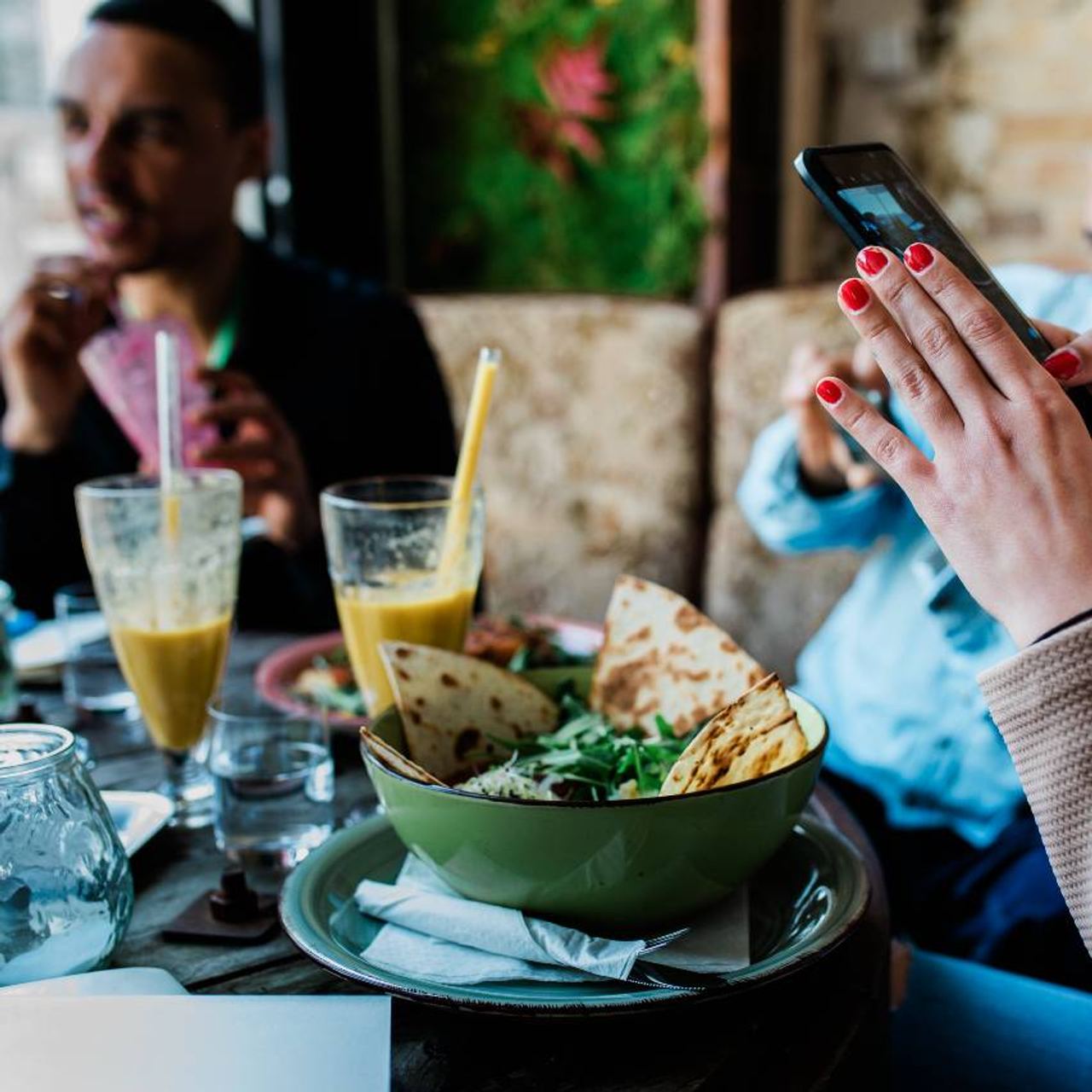 Menschen genießen gemeinsames Essen in einem gemütlichen Café, mit frischem Salat und Fladenbrot auf dem Tisch, während ein Gast ein Foto mit dem Smartphone macht.