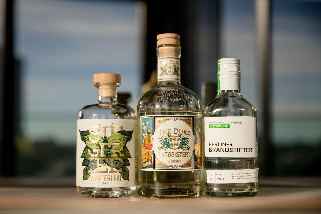 Drei verschiedene Gin-Flaschen mit Etiketten, stehend nebeneinander auf einem Holztisch, vor unscharfem Hintergrund mit Fenstern und Tageslicht.