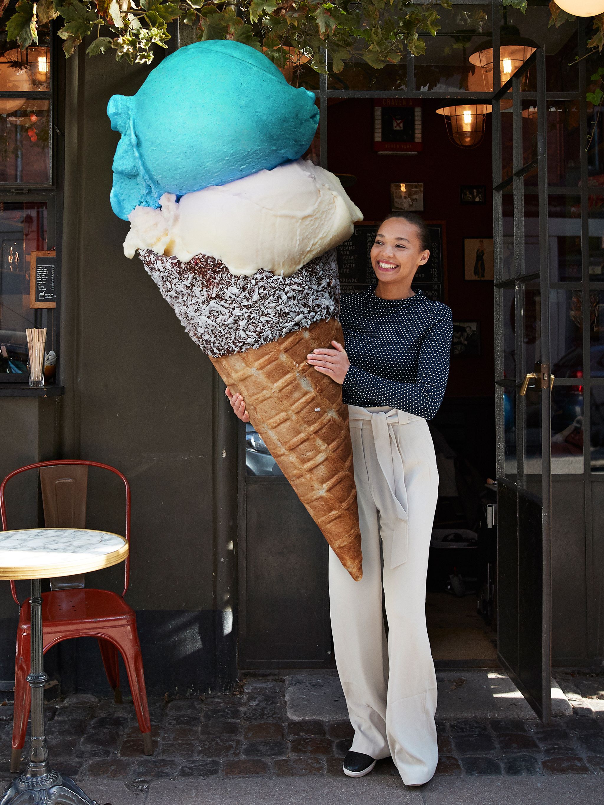 Eine Frau steht lächelnd vor einem Gebäude und hält eine überdimensionale Eiswaffel mit drei Kugeln in Blau, Weiß und Braun.
