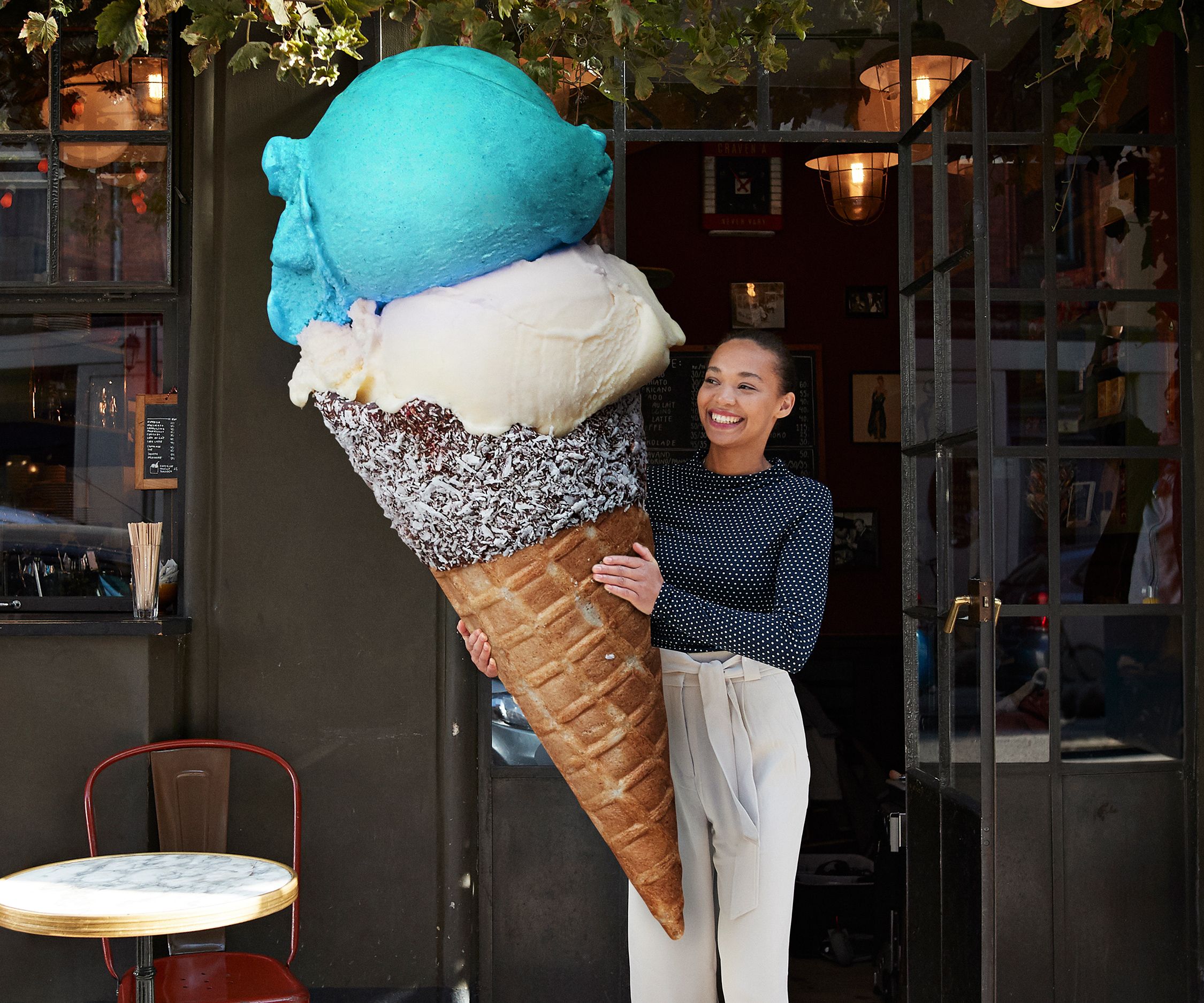Eine Frau steht lächelnd vor einem Eiscafé und hält eine übergroße Eistüte mit drei Kugeln in Blau, Weiß und Braun.