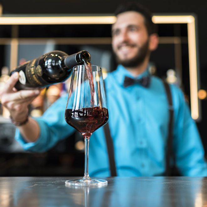 Ein Barkeeper in blauem Hemd füllt ein Glas mit rotem Wein, der Fokus liegt auf dem Glas, das stilvoll eingeschenkt wird.