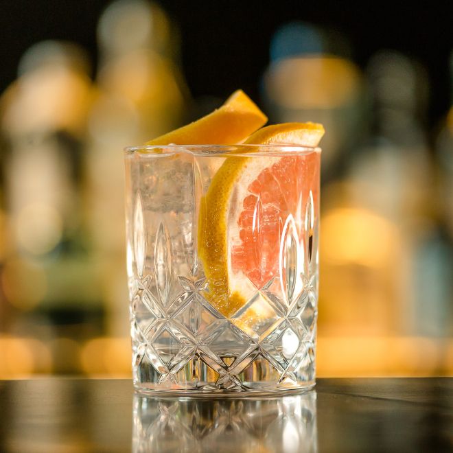 Klares Gin Getränk mit Eiswürfeln und einer Scheibe Grapefruit in einem dekorativen Kristallglas, abgestellt auf einer reflektierenden Oberfläche vor unscharf beleuchtetem Hintergrund.