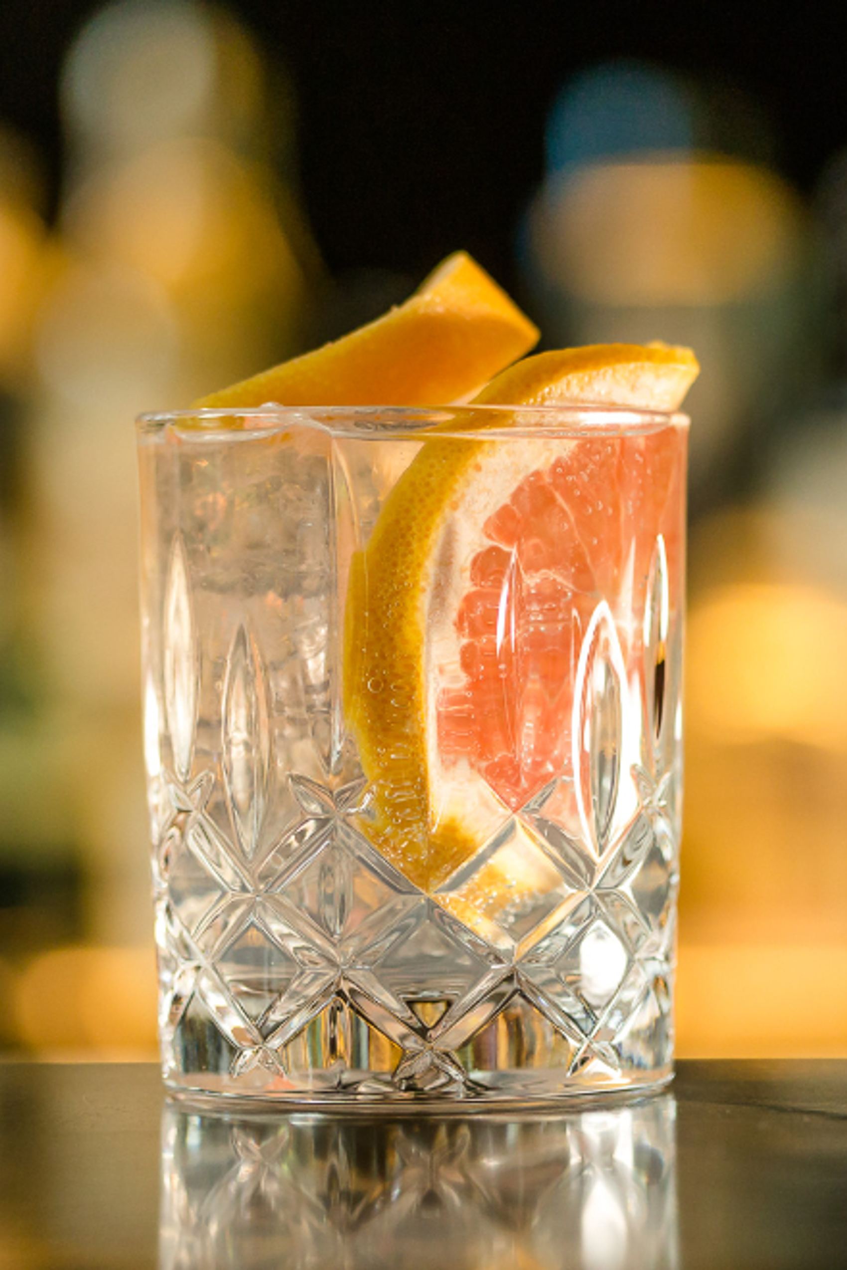 Klares Gin Getränk mit Eiswürfeln und einer Scheibe Grapefruit in einem dekorativen Kristallglas, abgestellt auf einer reflektierenden Oberfläche vor unscharf beleuchtetem Hintergrund.