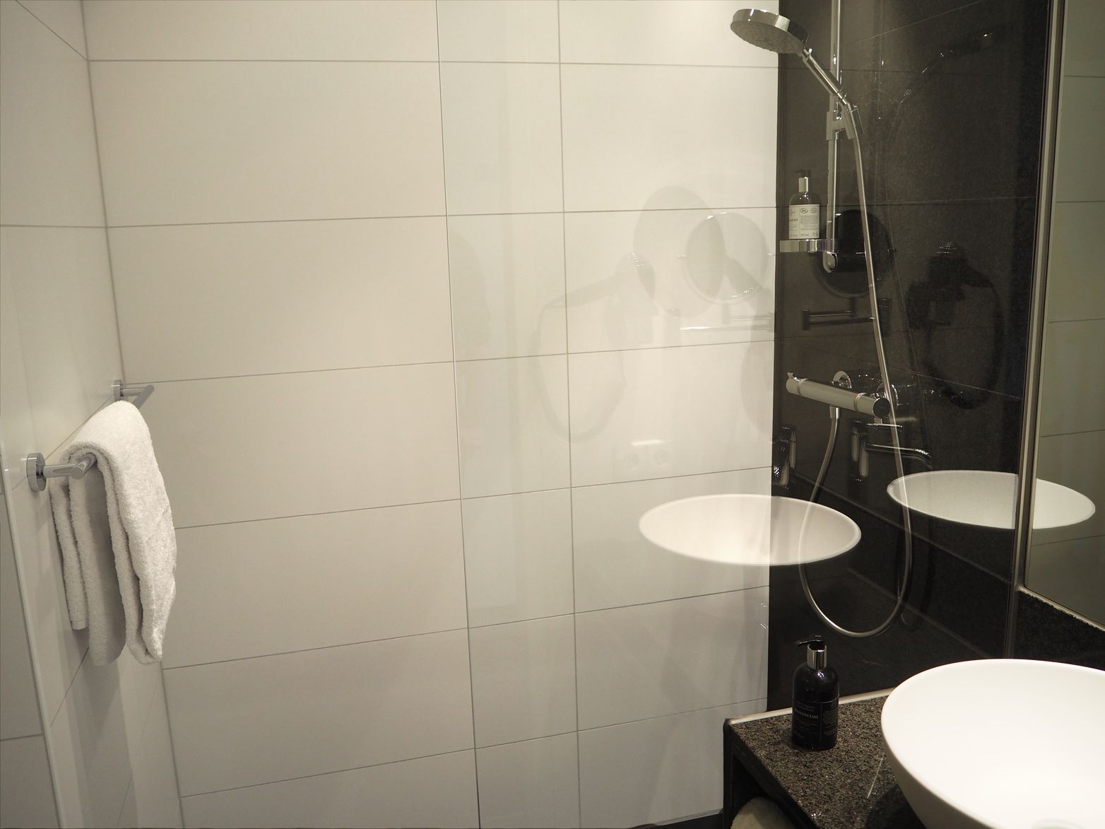 Badezimmer mit weißen Wandfliesen, rundes Waschbecken auf schwarzer Arbeitsfläche und Dusche mit Glasabtrennung. Ein weißes Handtuch hängt an einer Stange an der Wand.