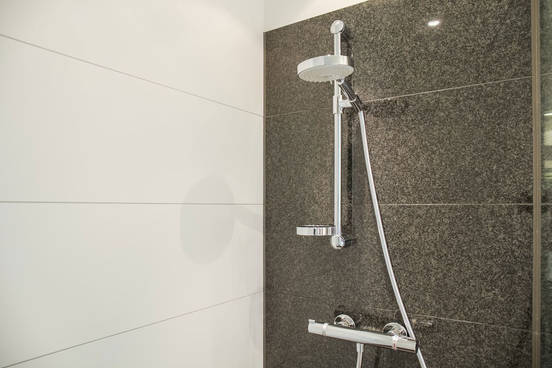 Moderne Dusche mit verstellbarem Duschkopf an dunkelgrauer Fliesenwand, weiße Fliesen an angrenzender Wand, minimalistisches und sauberes Design.