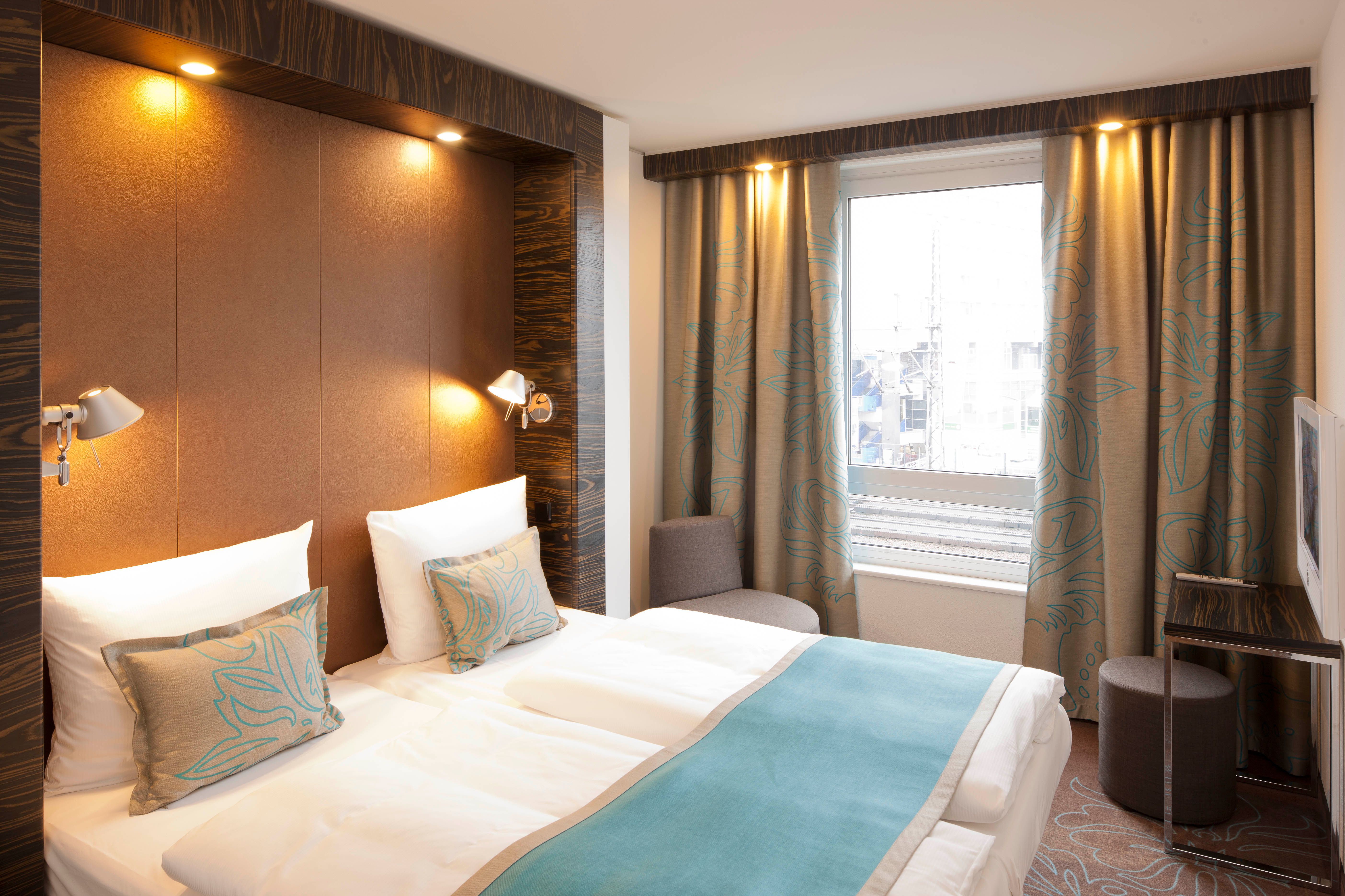 Motel One Hotel Zmmer mit Doppelbett mit zwei Matratzen, gemütlicher Beleuchtung, weichen Kissen und einem großen Fenster mit Vorhängen, das Tageslicht hereinlässt. Angenehme, ruhige Atmosphäre für erholsamen Aufenthalt.