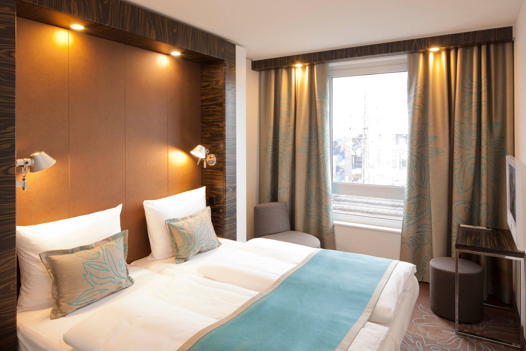 Motel One Hotel Zmmer mit Doppelbett mit zwei Matratzen, gemütlicher Beleuchtung, weichen Kissen und einem großen Fenster mit Vorhängen, das Tageslicht hereinlässt. Angenehme, ruhige Atmosphäre für erholsamen Aufenthalt.