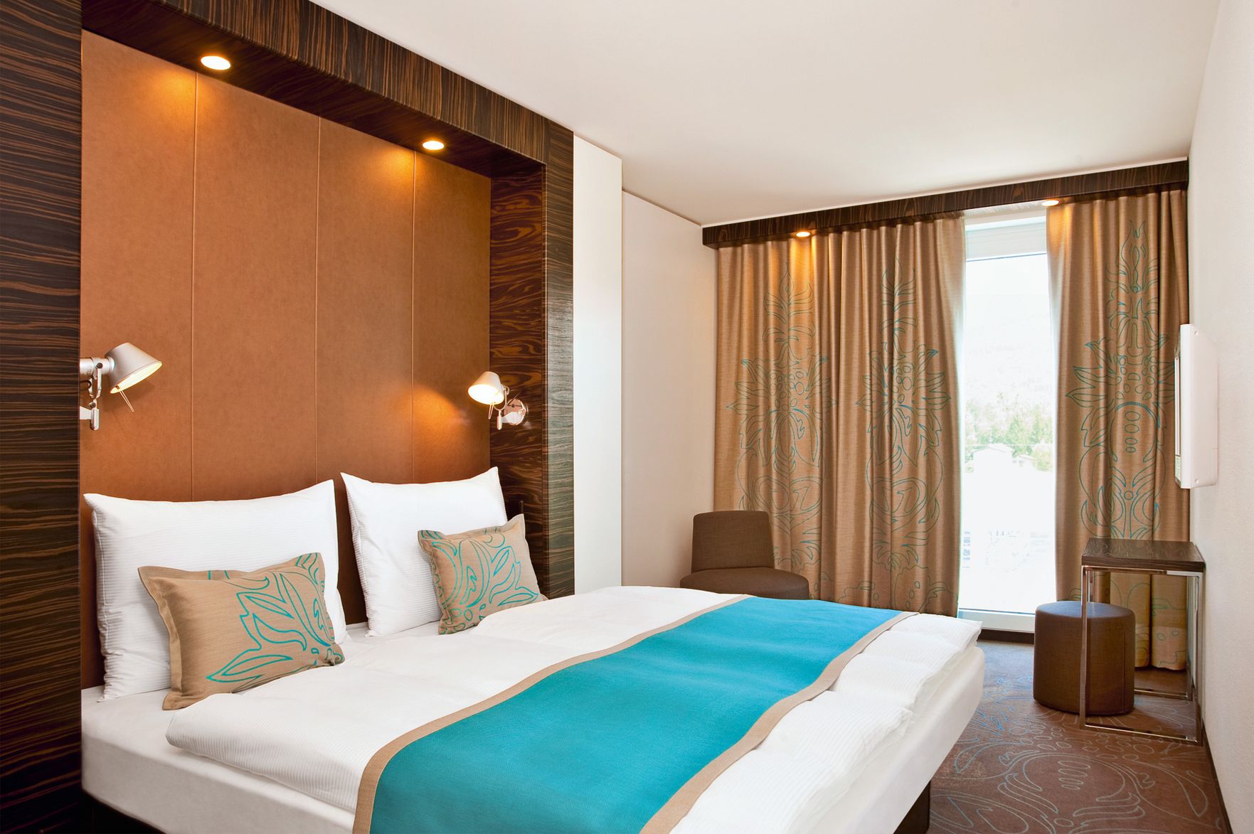 Motel One Hotel gemütliches Doppelzimmer mit großem Bett, hellen Wänden, warmem Licht, türkisem Bettläufer und dekorativen Kissen, sowie blickdichten Vorhängen vor einem Fenster mit Tageslicht.
