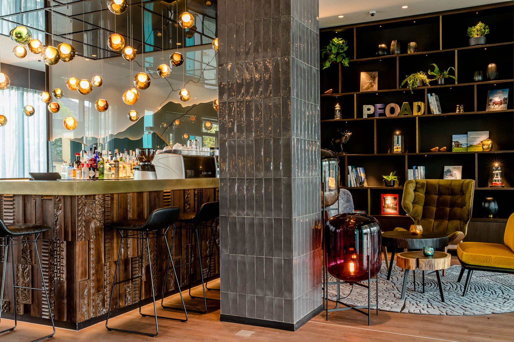 Elegante Hotelbar mit modernen Holz- und Metallakzenten, Barhockern und warmen Hängeleuchten. Gemütliche Lounge-Ecke mit Sesseln, Teppich und dekorativ beleuchtetem Bücherregal schafft entspannte Atmosphäre.