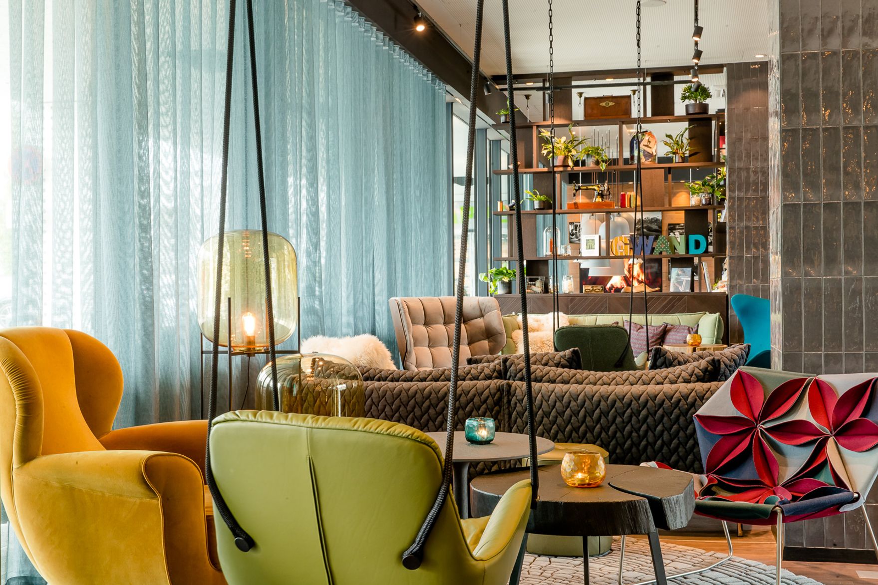 Helle, moderne Lounge mit bequemen Sesseln in verschiedenen Farben, einem großen Sofa und dekorativen Pflanzen, die eine einladende und entspannte Atmosphäre schaffen. Große Fenster mit lichtdurchlässigen Vorhängen sorgen für angenehmes Tageslicht.