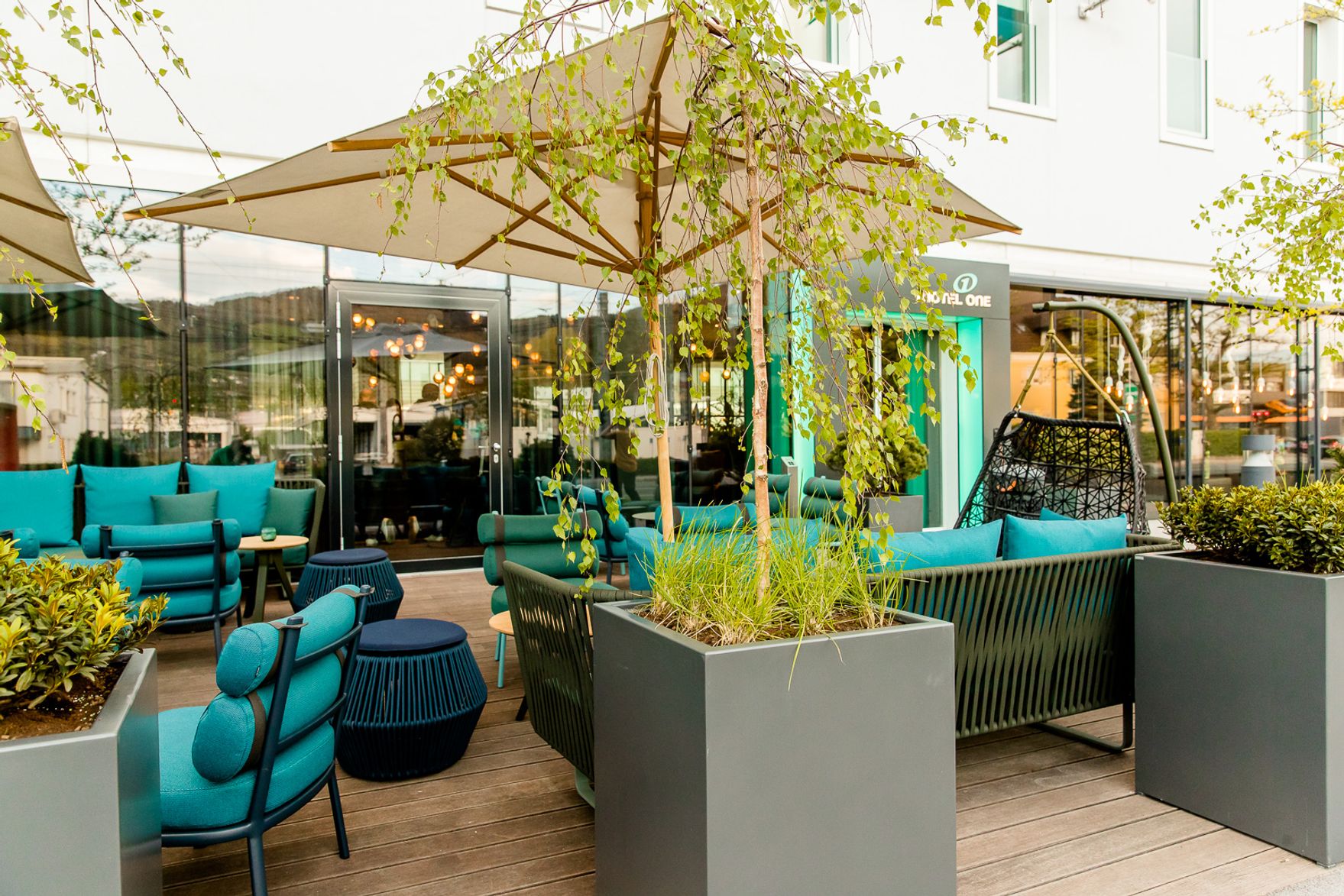 Outdoor-Loungebereich mit modernen blauen Sitzmöbeln, Sonnenschirm und großen Pflanzkübeln vor einem Hotelgebäude, ideal zum Entspannen und Genießen.
