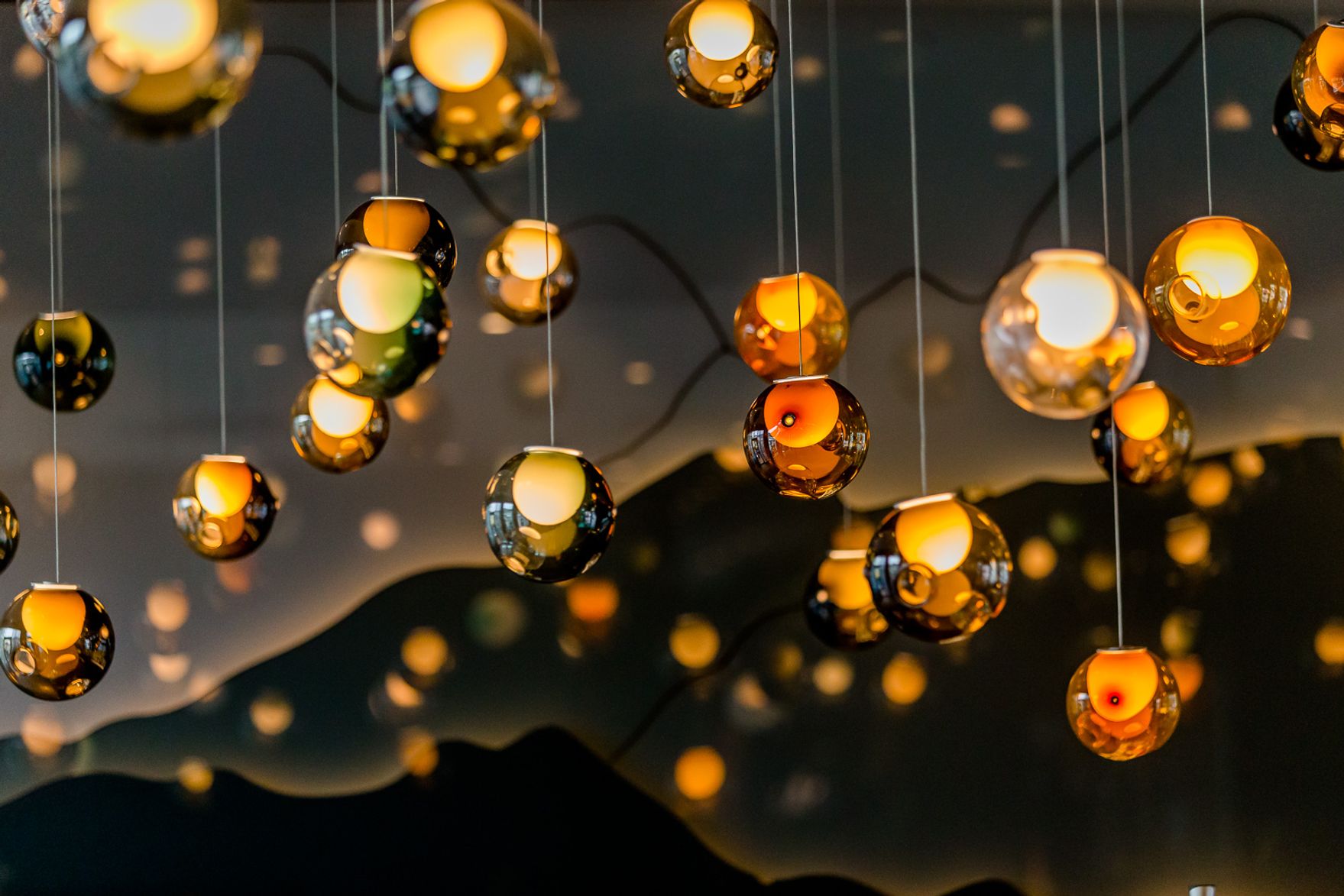 Kugelrunde, warm leuchtende Pendelleuchten hängen in unterschiedlicher Höhe an dünnen Kabeln, schaffen eine elegante und gemütliche Atmosphäre mit sanften Lichtreflexionen.