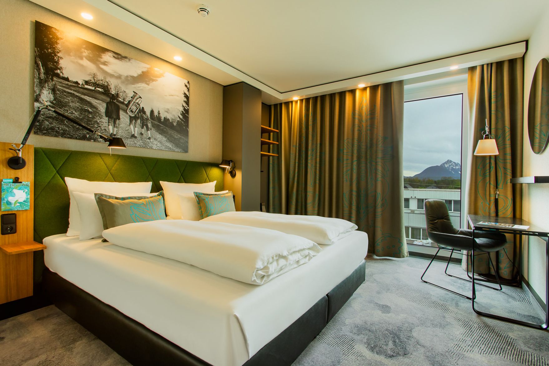 Modernes Hotelzimmer mit großem, komfortablem Doppelbett, stilvoller Beleuchtung und großem Fenster mit Blick auf Berge. Elegantes Ambiente mit schlichten, warmen Farben und funktionalem Arbeitsplatz.