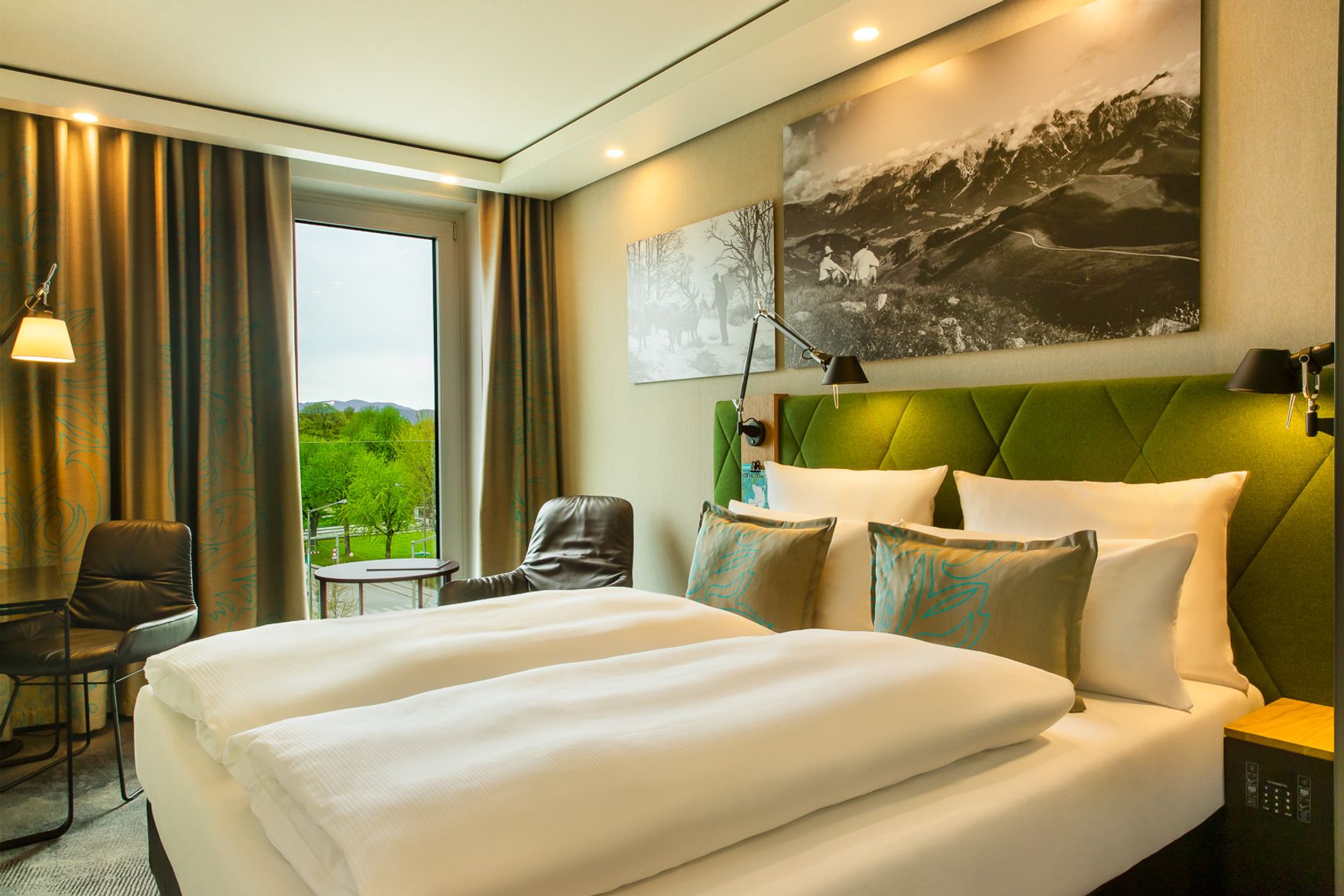 Modernes Hotelzimmer mit großem Doppelbett, grüner Polsterung am Kopfteil, Blick auf grüne Landschaft durch großes Fenster, gemütlicher Sitzbereich mit zwei Sesseln und warmem, einladendem Ambiente.