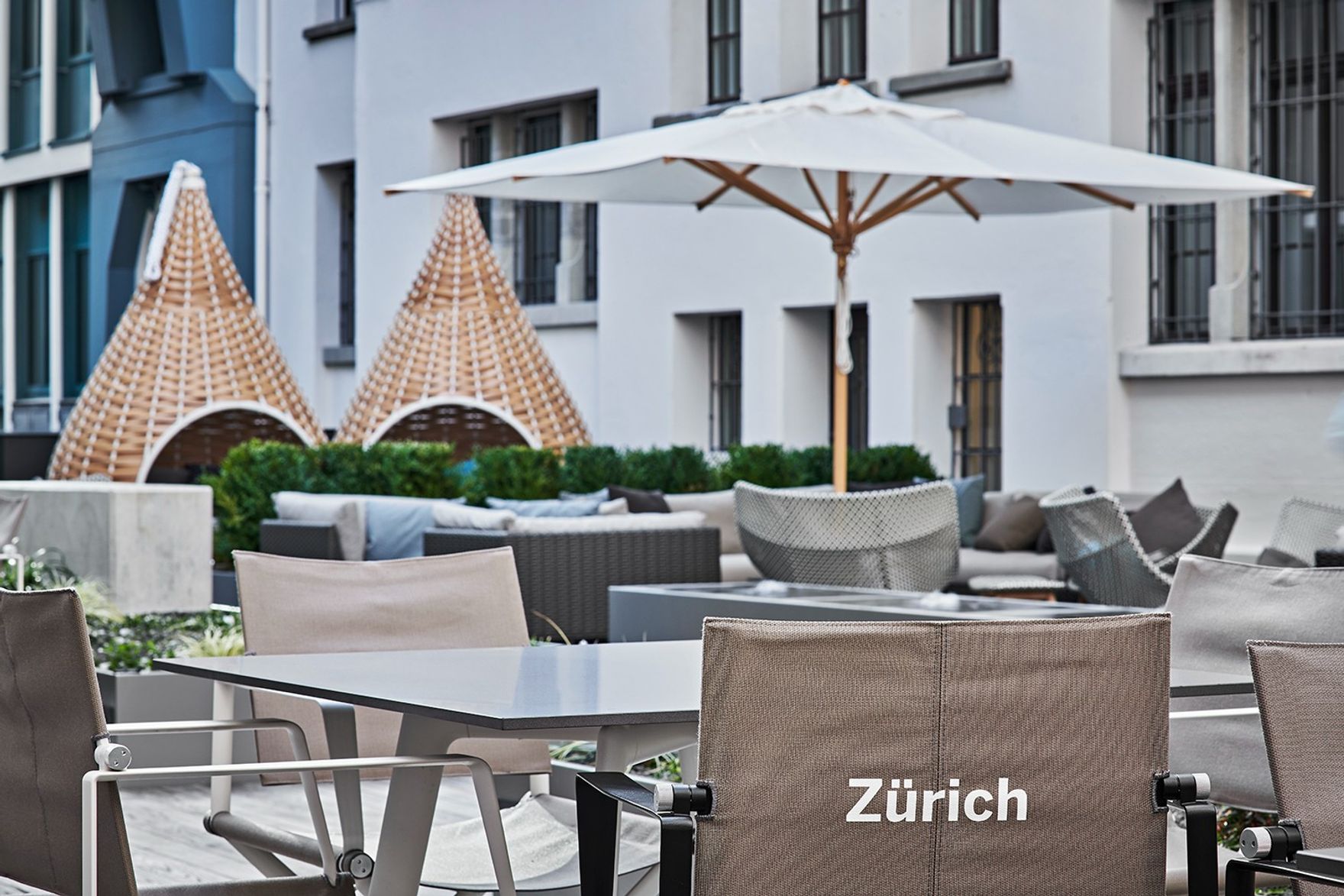Außenterrasse mit komfortablen Sitzmöbeln, Sonnenschirm und dekorativen, geflochtenen Lounge-Hängesesseln vor einem modernen Gebäude, ideal für entspannte Stunden in Zürich.