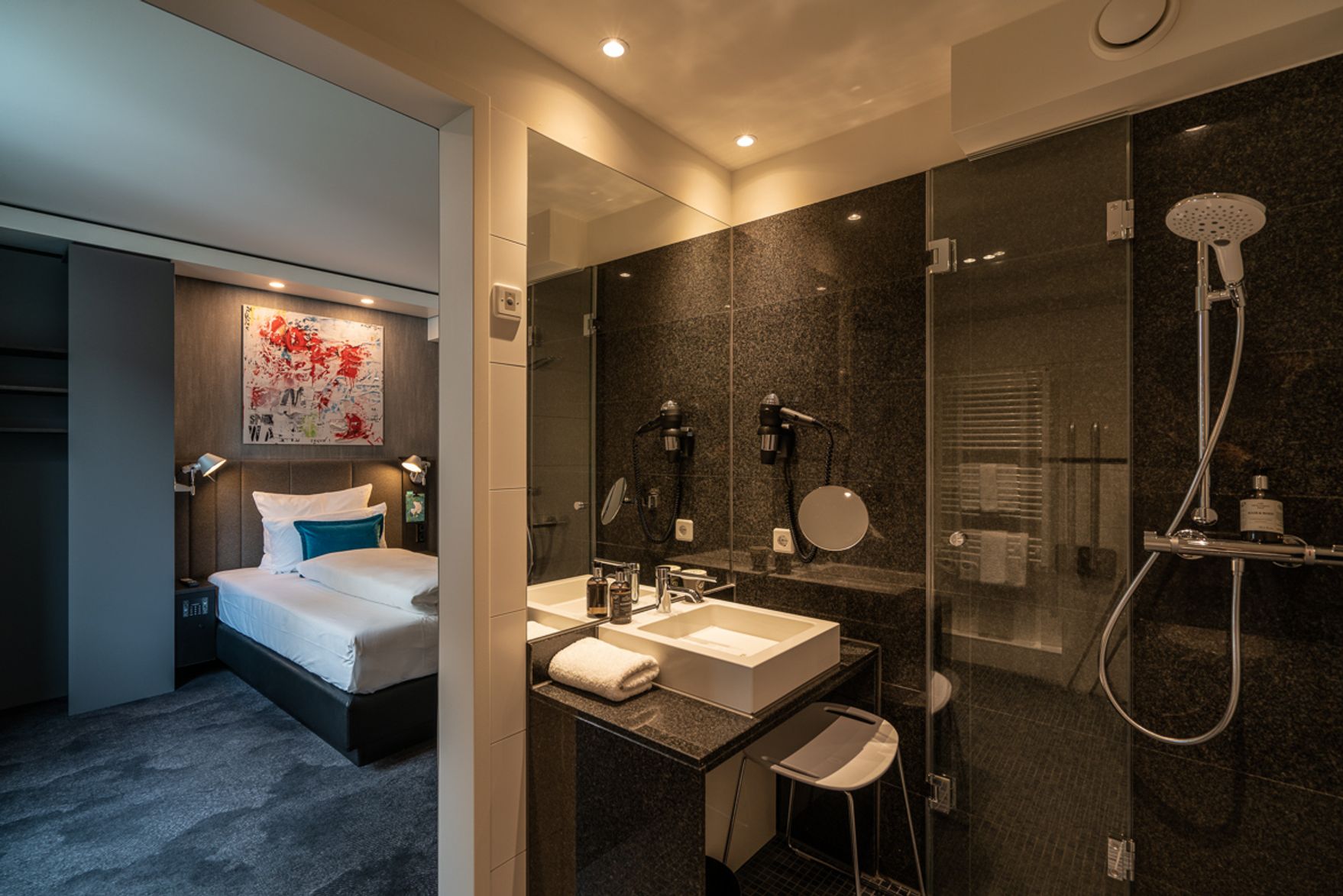 Modernes Hotelzimmer mit komfortablem Einzelbett, stimmungsvoller Beleuchtung und farbenfroher Wandkunst, angrenzendem Badezimmer mit Dusche, Waschbecken und Haartrockner – ideal für Geschäftsreisende und Erholungssuchende.
