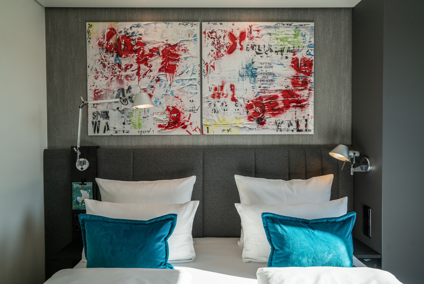 Modern eingerichtetes Doppelzimmer mit großem, gepolstertem Kopfteil, weißer Bettwäsche und leuchtend blauen Kissen. Über dem Bett hängen zwei abstrakte, farbenfrohe Gemälde, die dem Raum eine lebendige Atmosphäre verleihen. Sanfte Beleuchtung durch Leselampen an beiden Seiten des Bettes.