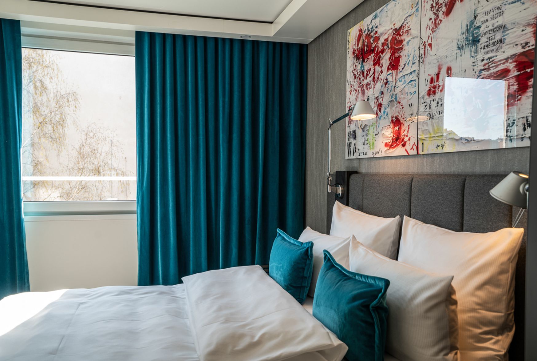 Modern eingerichtetes Hotelzimmer mit großem Bett, weißer Bettwäsche, türkisfarbenen Vorhängen und passenden Kissen. Kunstvoll gestaltetes Wandbild über dem Kopfteil sorgt für stilvolle Atmosphäre. Natürliches Tageslicht durch Fenster.