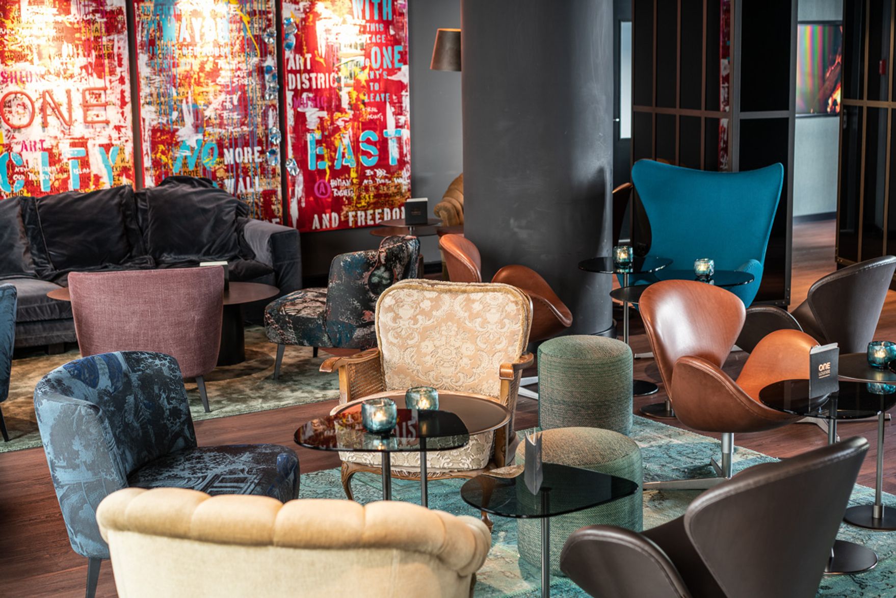 Moderne Hotel-Lounge mit verschiedenen bequemen Sesseln und Sofas in unterschiedlichen Farben, kleinen Glastischen und einer bunten, künstlerischen Wand im Hintergrund, die eine entspannte und einladende Atmosphäre für Gäste schafft.