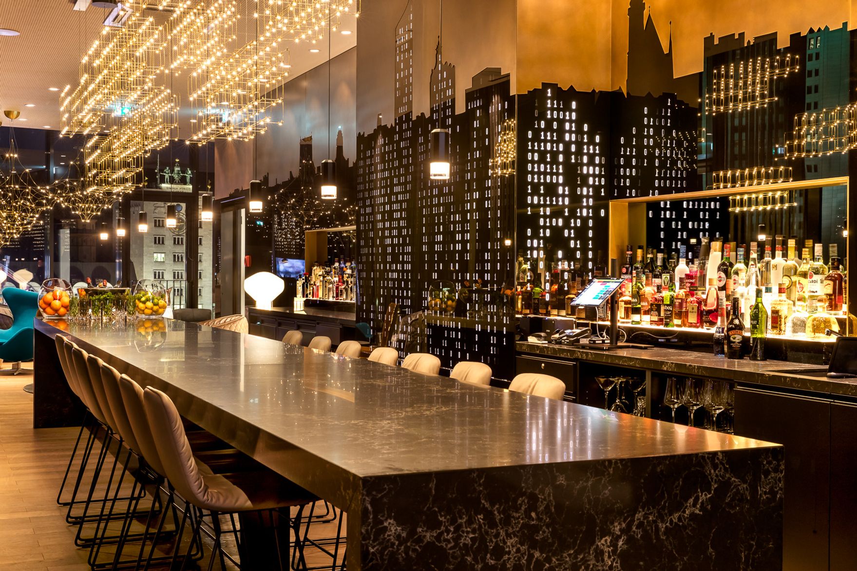 Hotel Leipzig Post Motel One Bar mit langer Marmortheke, eleganten Barhockern und stimmungsvoller Beleuchtung. Wanddekor zeigt stilisierte Stadtsilhouette bei Nacht, schafft urbane, gemütliche Atmosphäre für entspannte Abende und geselliges Beisammensein.