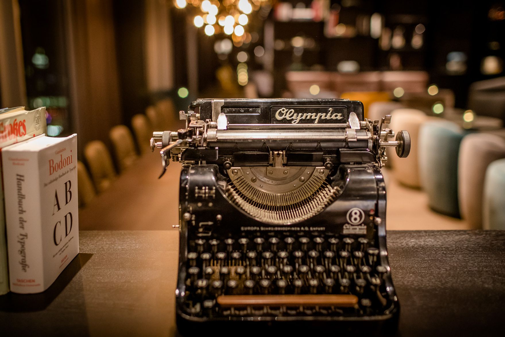 Eine antike schwarze Schreibmaschine „Olympia“ steht im Fokus, umgeben von Büchern und warm beleuchtetem Ambiente, das an eine gemütliche Lese- oder Arbeitsecke in einem stilvollen Hotel erinnert.