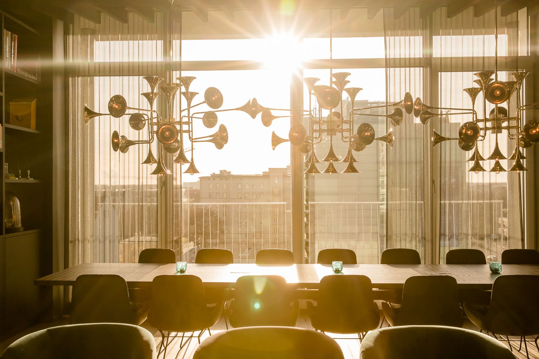 Sonnenlicht strömt durch bodentiefe Fenster in einen modernen Speiseraum mit langem Tisch und schwarzem Stühlen, klare, luftige Atmosphäre mit warmem Licht und dekorativen, goldfarbenen Hängeleuchten.