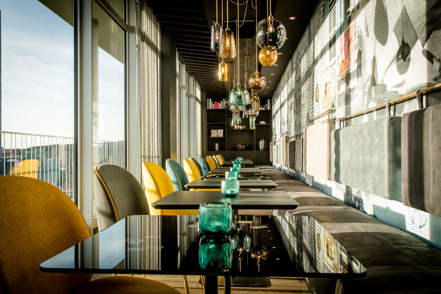 Modernes Restaurant mit elegantem Interieur, großen Fenstern und gemütlichen Stühlen in Gelb und Grau, stilvollen Hängeleuchten und Tischen mit grünen Gläsern, die ein warmes, einladendes Ambiente und Ausblick auf die Landschaft bieten.