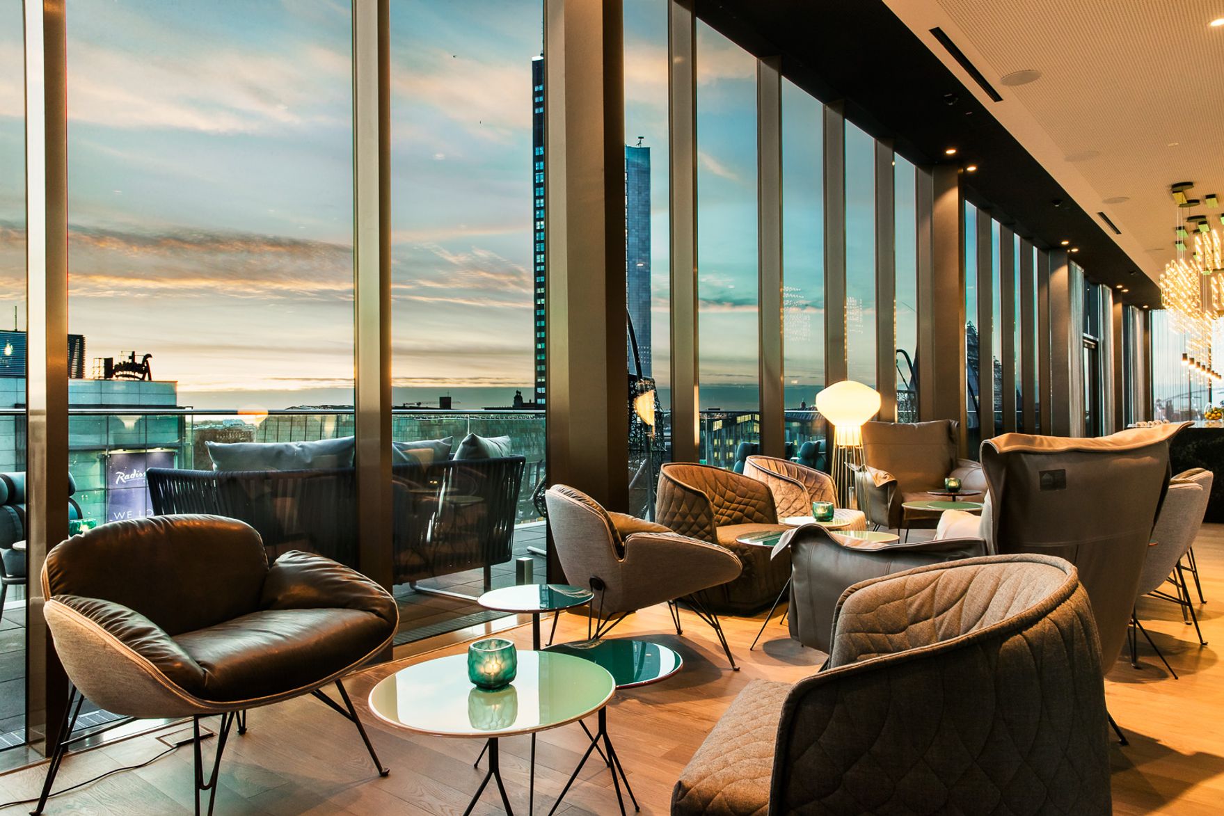Gemütliche Lounge mit modernen Sesseln und Tischen, großer Fensterfront mit Blick auf den Sonnenuntergang und die Stadt, warme Beleuchtung für entspannte Atmosphäre.