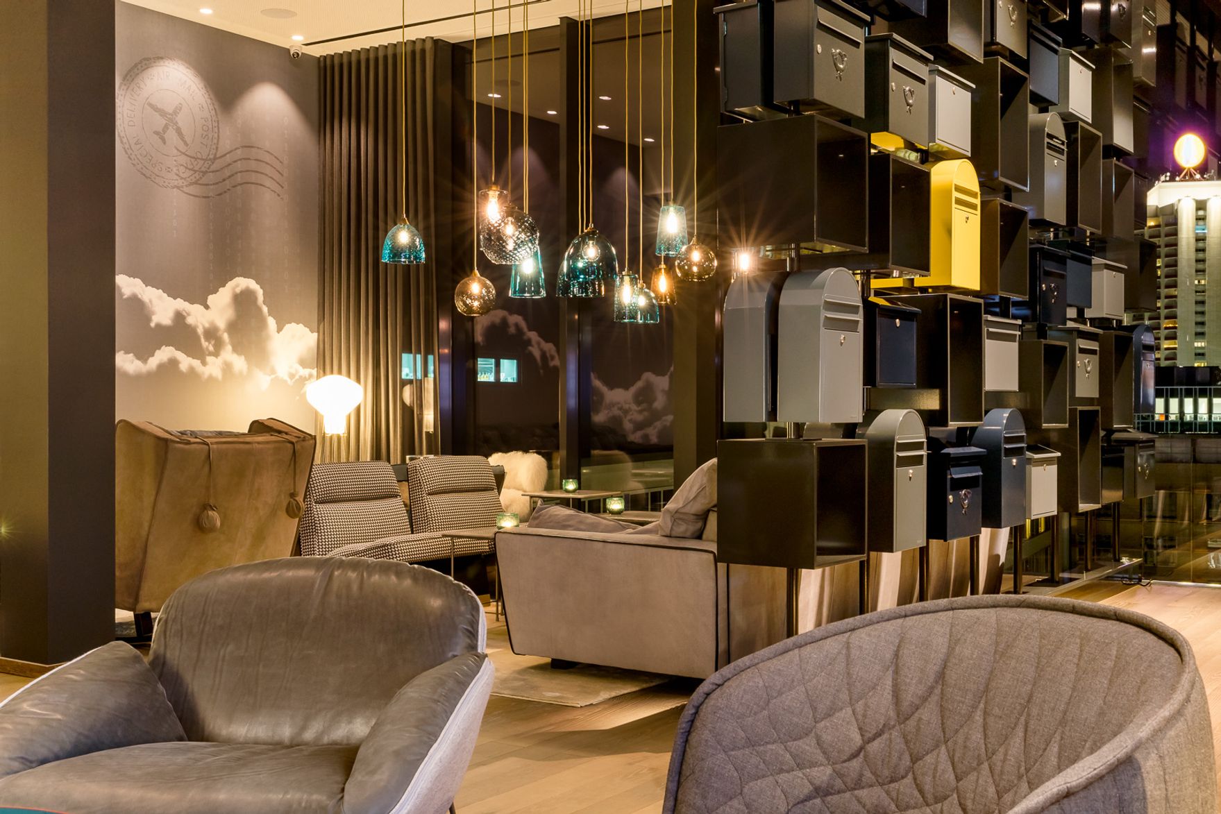 Gemütliche Hotellobby mit bequemen Sesseln und Sofas, stimmungsvoller Beleuchtung durch hängende Lampen und dekorativer Wandgestaltung mit Wolkenmotiv. Kreative Raumteiler aus verschiedenfarbigen Briefkästen schaffen eine moderne und einladende Atmosphäre zum Entspannen und Verweilen.