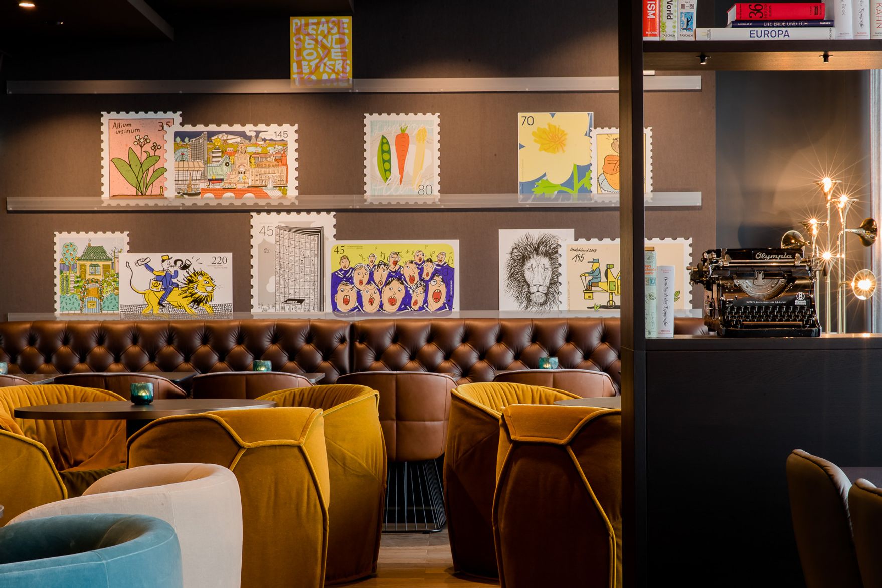 Gemütliche Hotel-Lounge mit braunem Ledersofa, senffarbenen Sesseln und kreativer Wanddekoration in Form großer Postkartenmotive, die eine warme, einladende Atmosphäre schaffen.