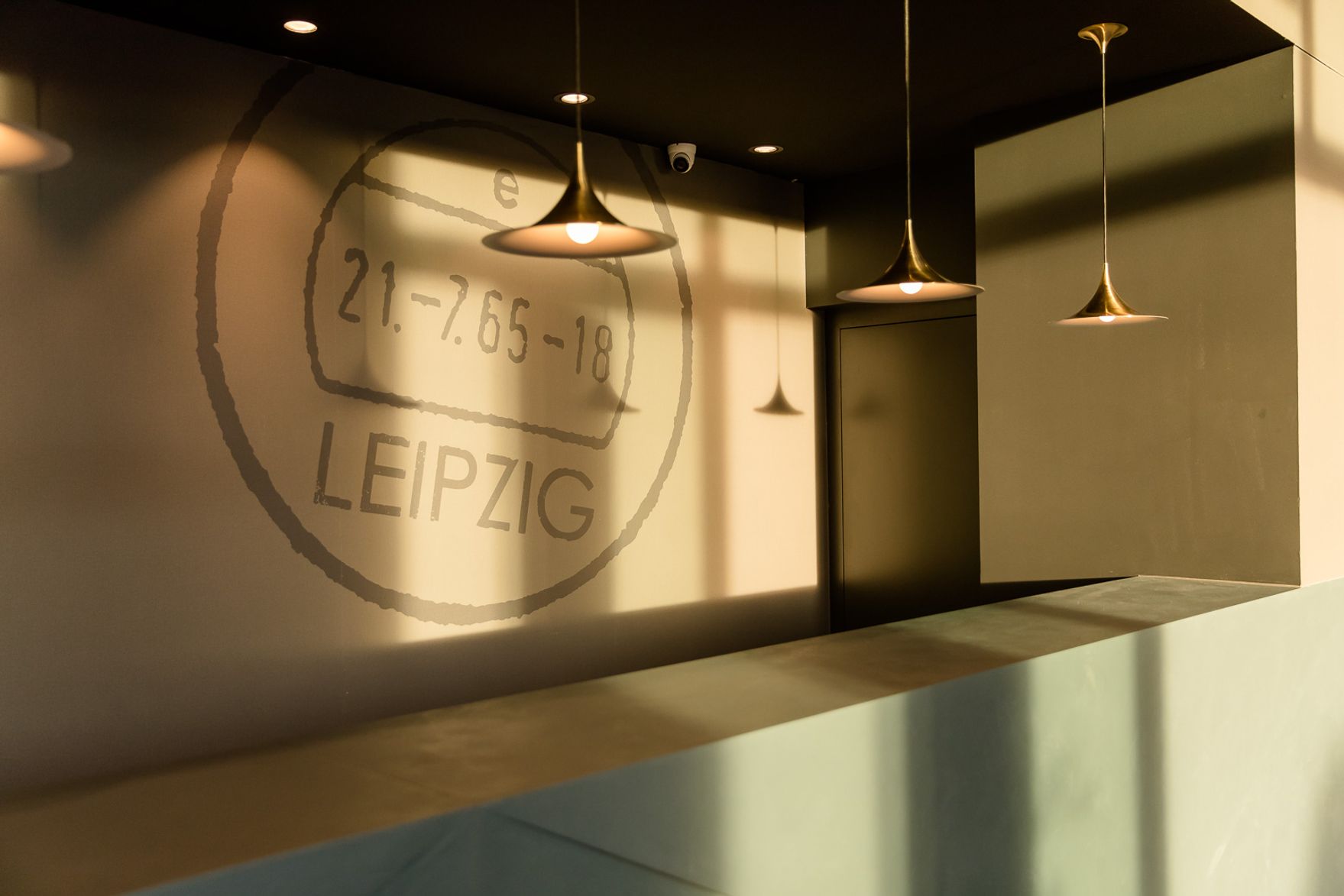 Empfangsbereich mit modernem, schlichtem Tresen und warmem Licht, das eine dekorative Projektion mit dem Schriftzug „Leipzig“ an die Wand wirft. Schlichte Pendelleuchten sorgen für eine einladende Atmosphäre.