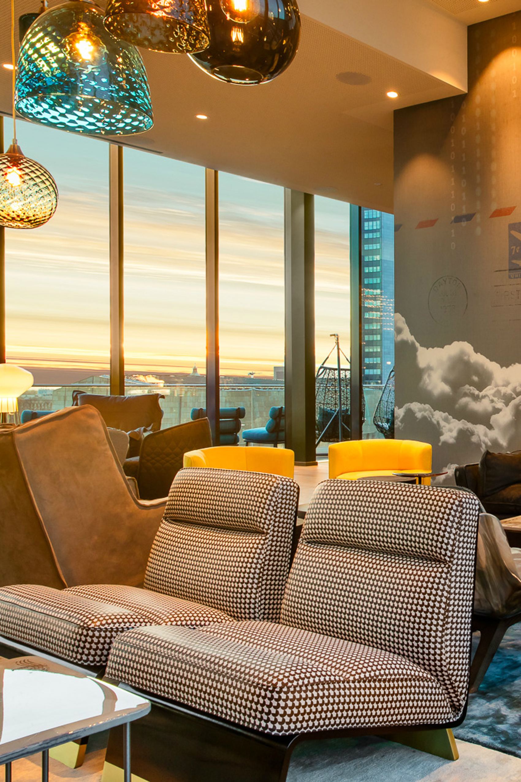 Gemütliche Lounge mit stilvollen Sesseln und warmem Licht, großer Fensterfront mit Blick auf den Sonnenuntergang über der Stadt. Moderne, einladende Atmosphäre ideal zum Entspannen und Genießen.