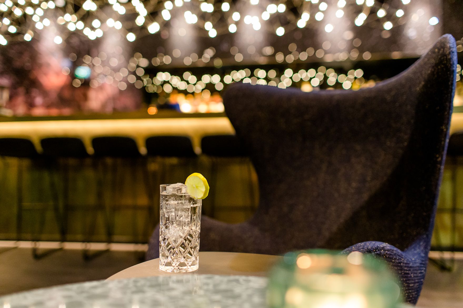 Glas mit erfrischendem Getränk und Zitronenscheibe auf Tisch vor gemütlichem Sessel, warm beleuchtete Bar im Hintergrund mit sanften Lichtern, elegante und entspannte Atmosphäre.